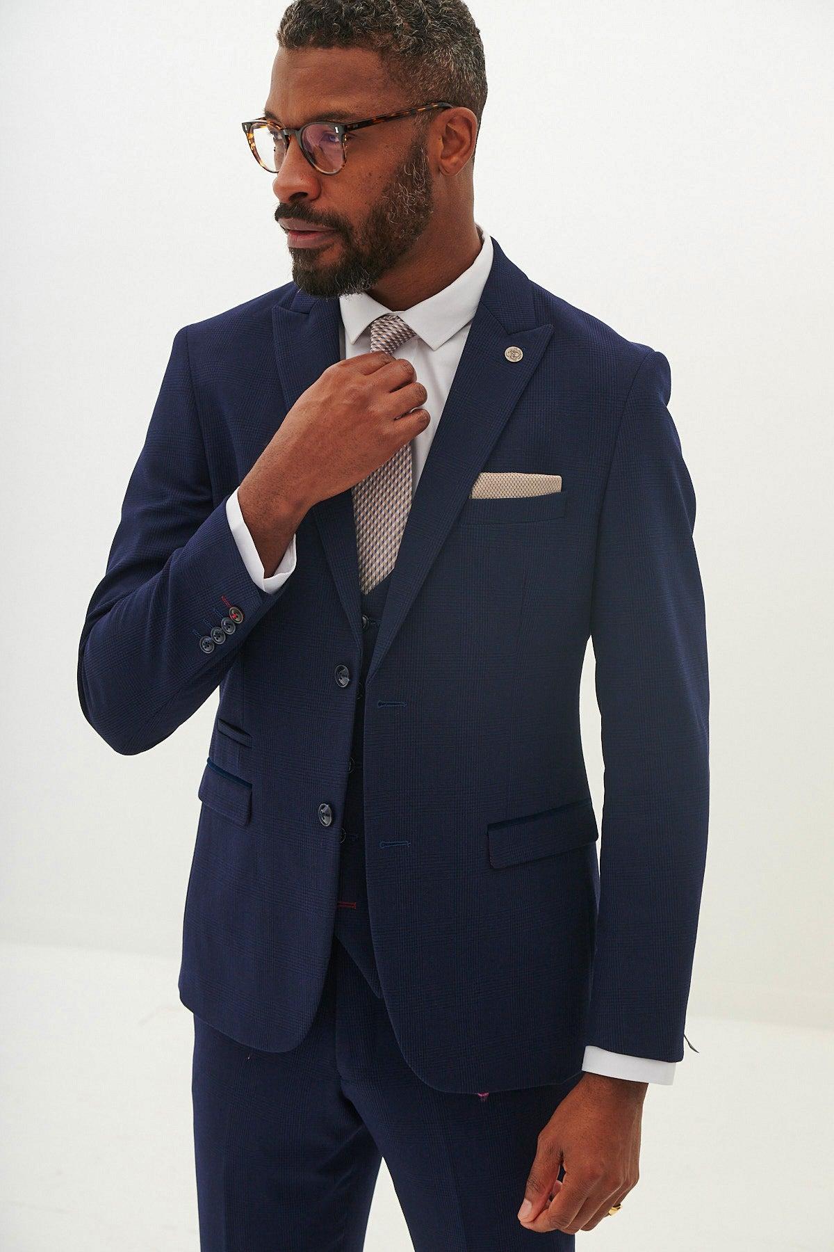 A man wears the Marc Darcy BROMLEY Navy Check Blazer with a white dress shirt and patterned tie, adjusting his tie and wearing glasses as he stands against a plain white background.