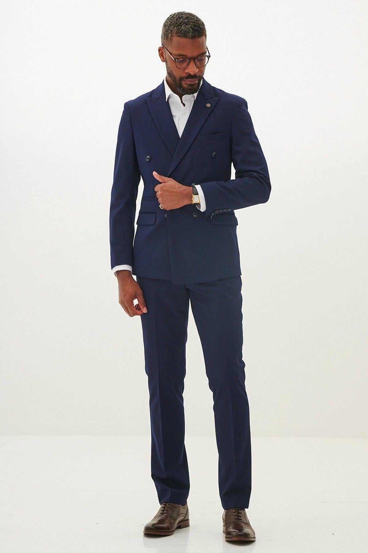 A man wears the Marc Darcy BROMLEY Navy Double Breasted Blazer with HM5 Stone Trousers, paired with a white shirt, brown shoes, and glasses. He stands against a white background with his hand on his jacket lapel.