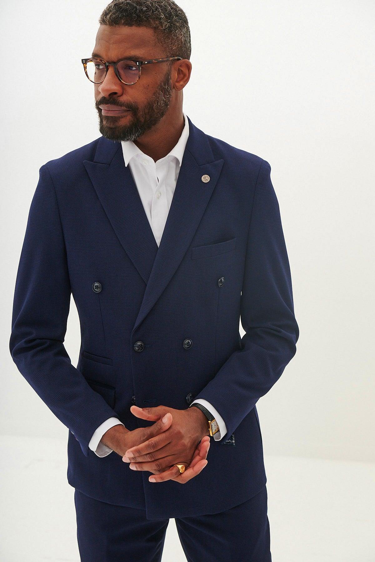 A man with short hair, a beard, and glasses wears the Marc Darcy BROMLEY Navy Double Breasted Check Blazer over a white shirt, standing against a plain light background with his hands clasped in front.