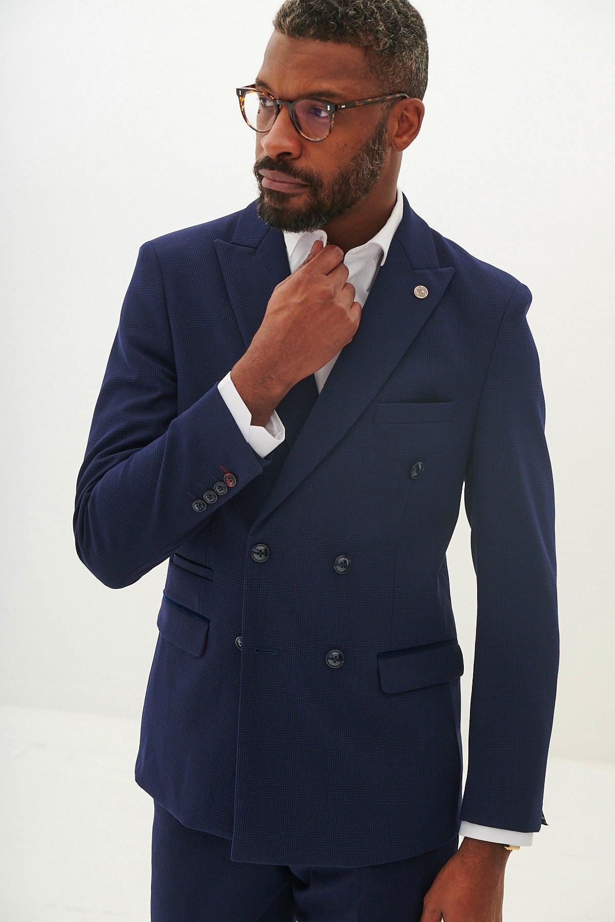A man with short hair, a beard, and glasses adjusts his collar while wearing the Marc Darcy BROMLEY Navy Double Breasted Blazer with HM5 Stone Trousers and a white shirt, looking to the side against a plain white background.