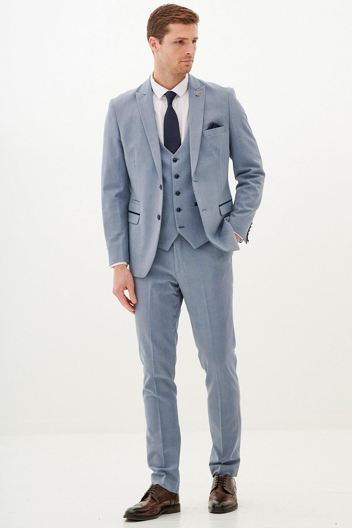 A man poses against a white background in the Marc Darcy BROMLEY - Sky Blue Check Three Piece Suit, styled with a white shirt, dark blue tie, and brown dress shoes. He stands with one hand by his side and gazes slightly to the side.