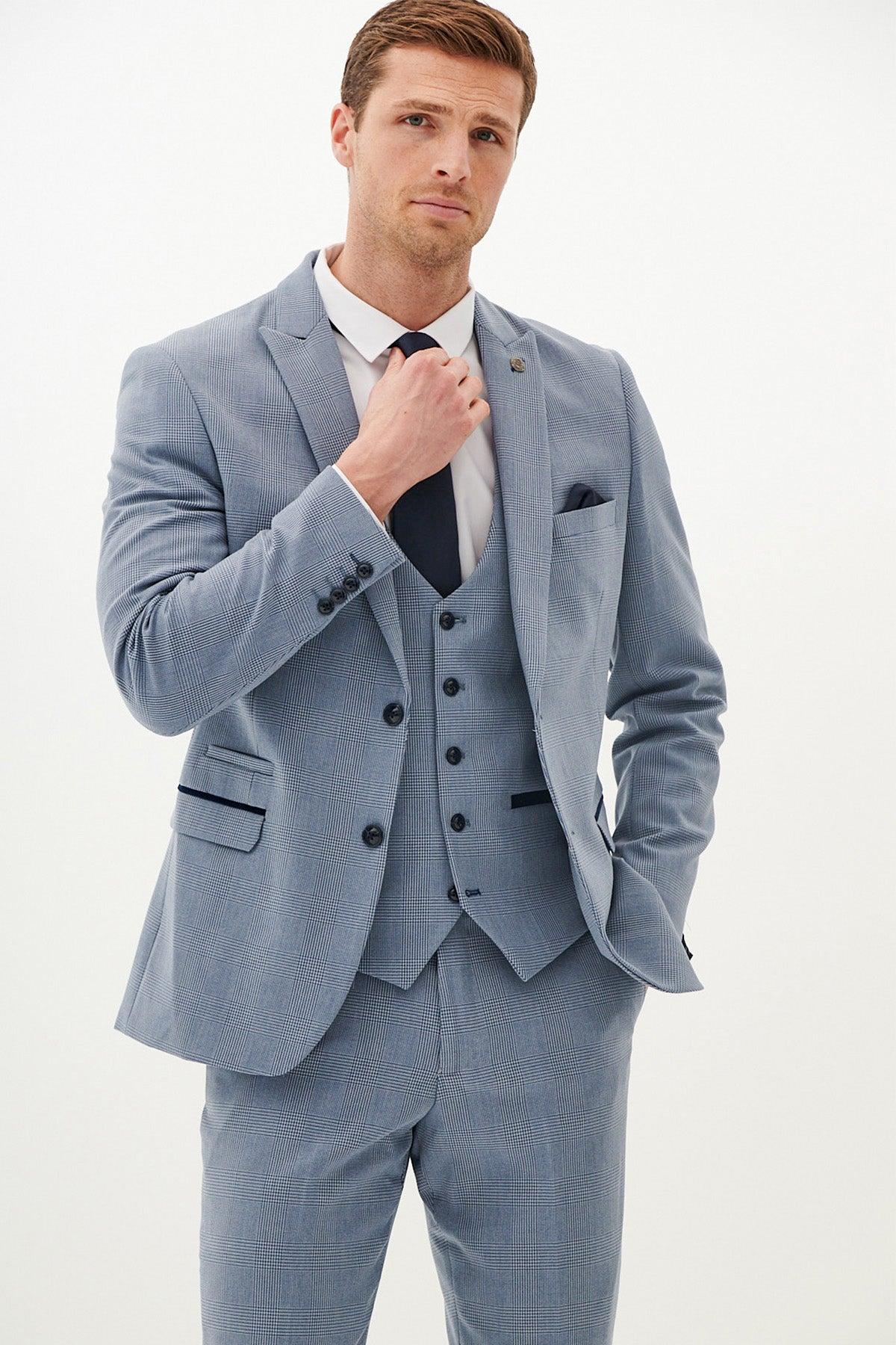 A man in the Marc Darcy BROMLEY Sky Blue Check Three Piece Suit with a navy tie adjusts his tie against a white background.