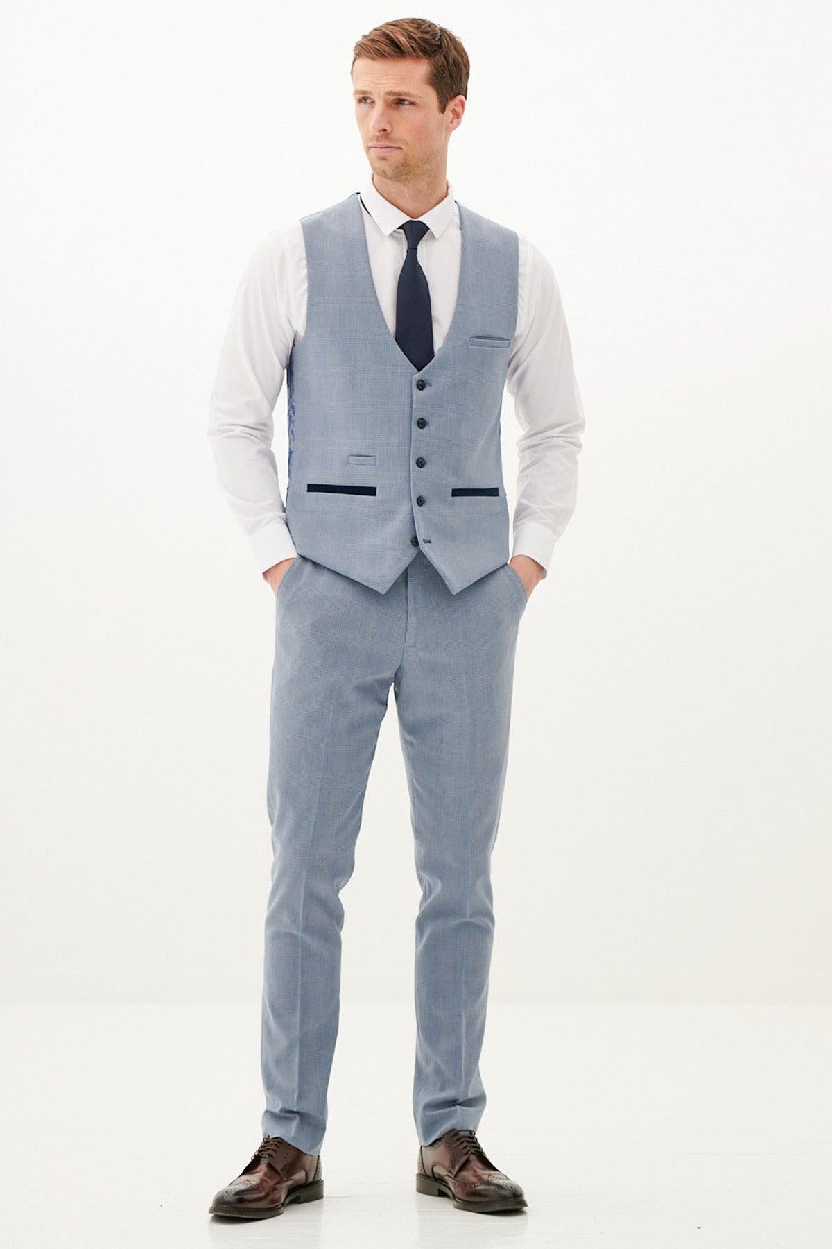 A man wears the Marc Darcy BROMLEY sky blue single breasted check waistcoat, paired with a white shirt and navy tie, standing with hands in his pockets against a white background.