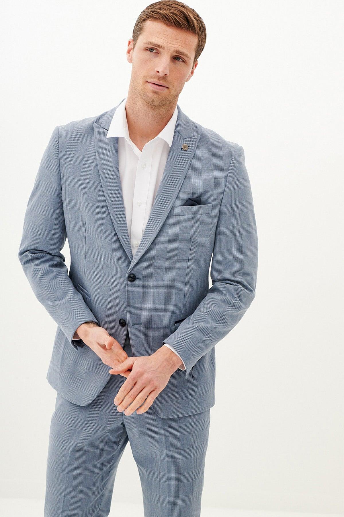 A man wears the Marc Darcy BROMLEY - Sky Blue Check Blazer over a white dress shirt, standing against a white background and looking at the camera with a relaxed expression and his hands together.