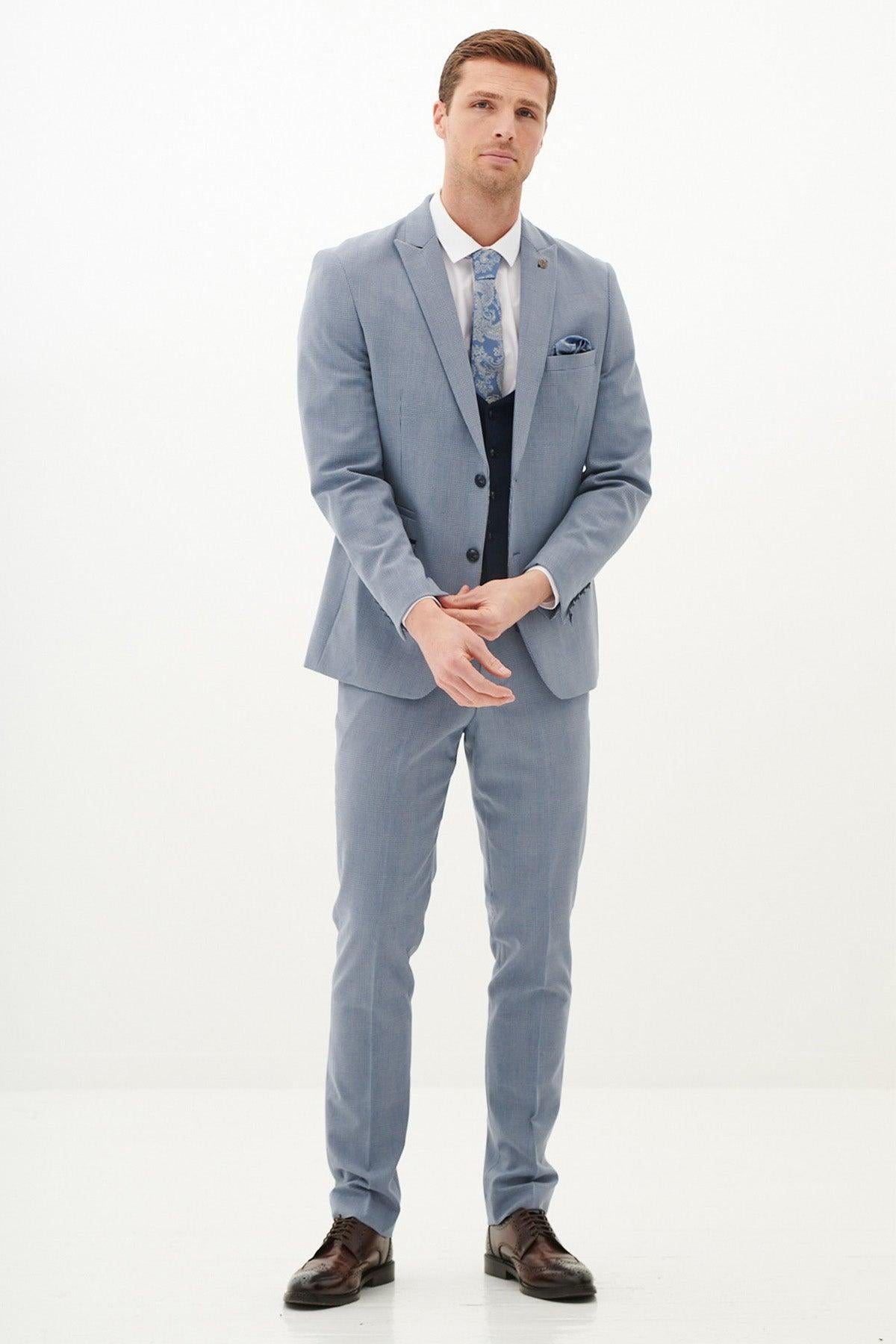 A man stands full-body in the Marc Darcy BROMLEY - Sky Check Suit with Kelvin Navy Waistcoat, white shirt, and patterned blue tie, looking at the camera.