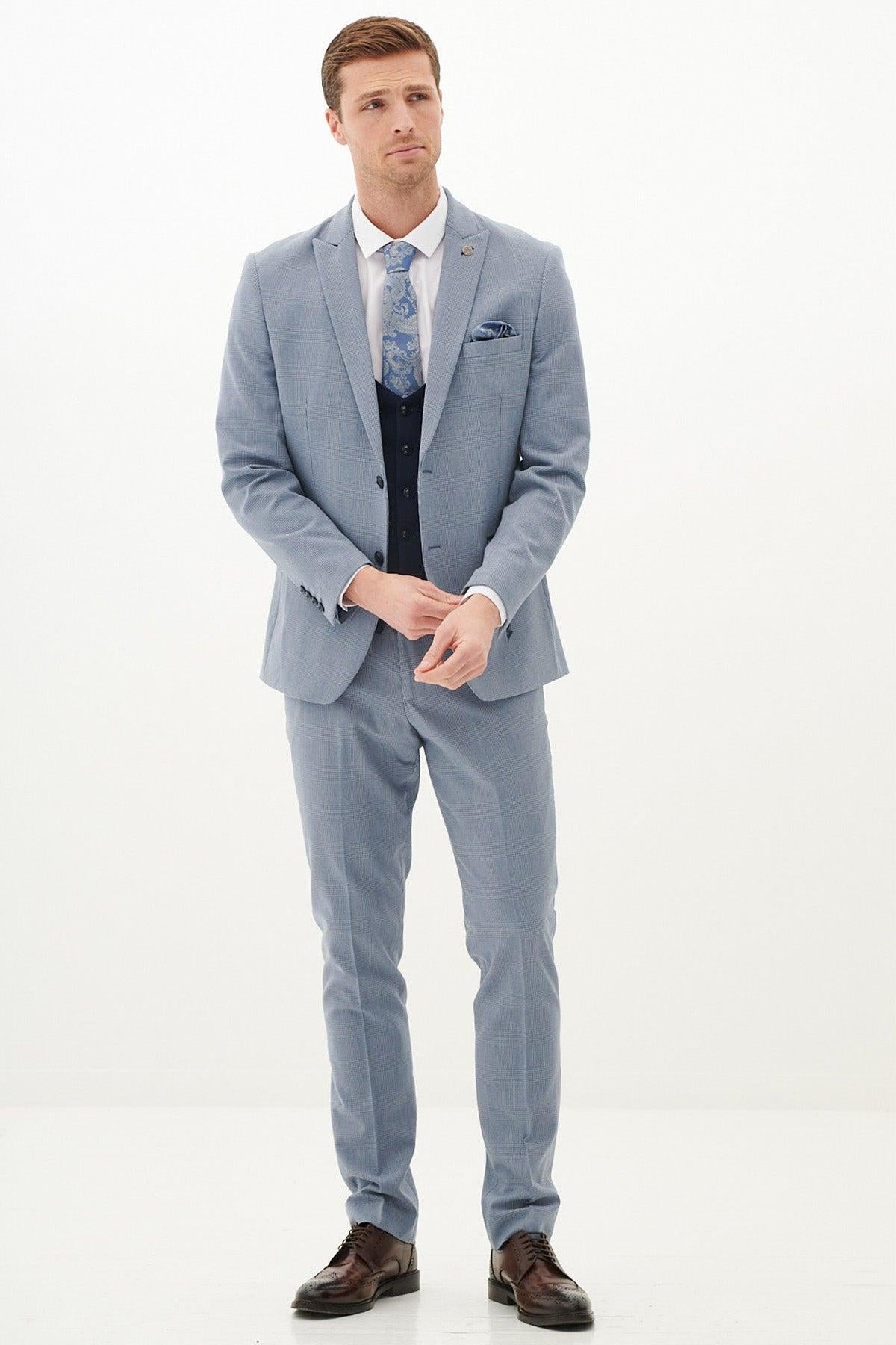 A man stands against a white background wearing the Marc Darcy BROMLEY Sky Check Suit with Kelvin Navy Waistcoat, white shirt, blue patterned tie, pocket square, and brown shoes. He looks slightly aside while adjusting his cuff.