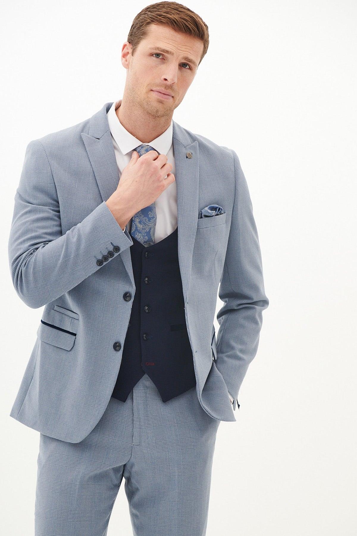 A man in a Marc Darcy BROMLEY Sky Check Suit with Kelvin Navy Waistcoat, white shirt, and blue patterned tie stands against a plain white background, adjusting his tie with one hand and keeping the other in his pocket.