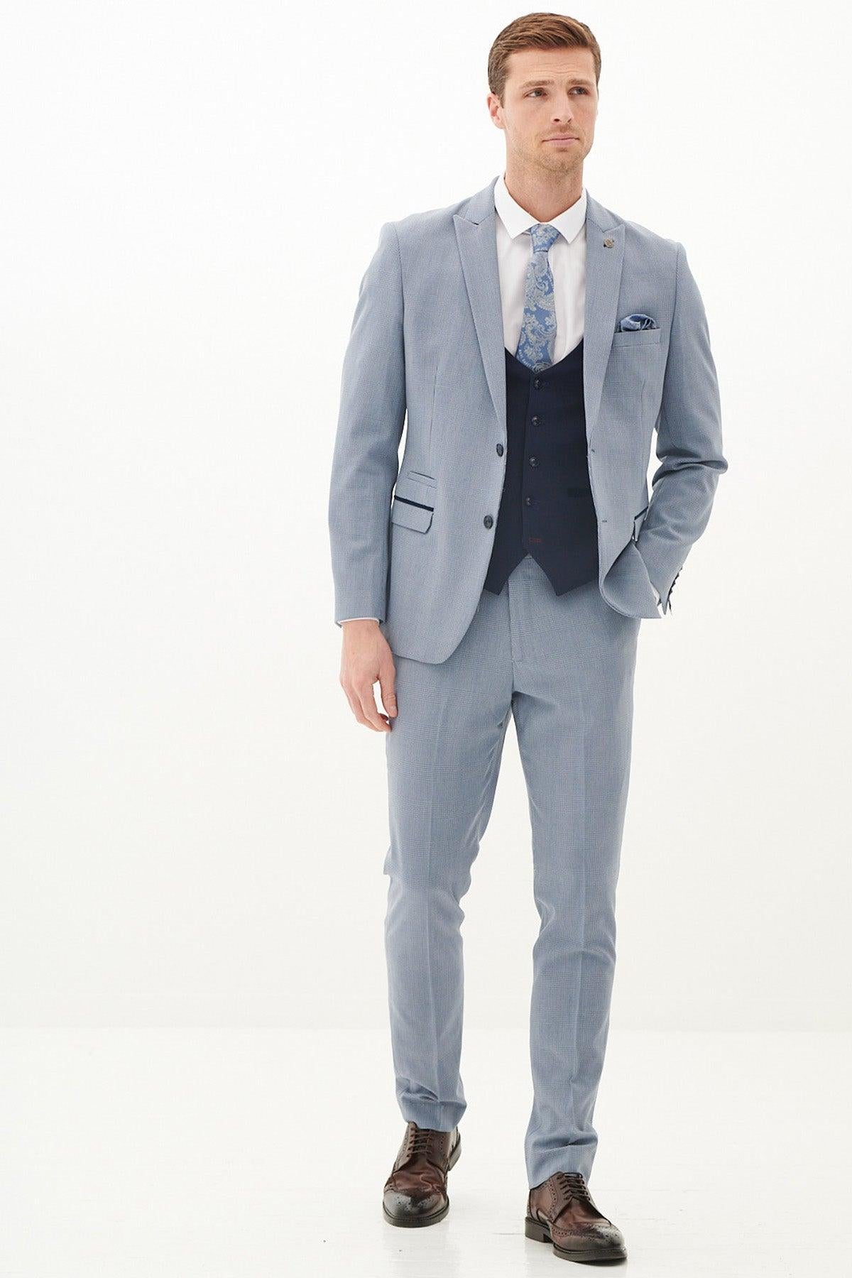 A man in a Marc Darcy BROMLEY - Sky Check Suit with Kelvin Navy Waistcoat, white shirt, patterned tie, and brown dress shoes stands against a white background.