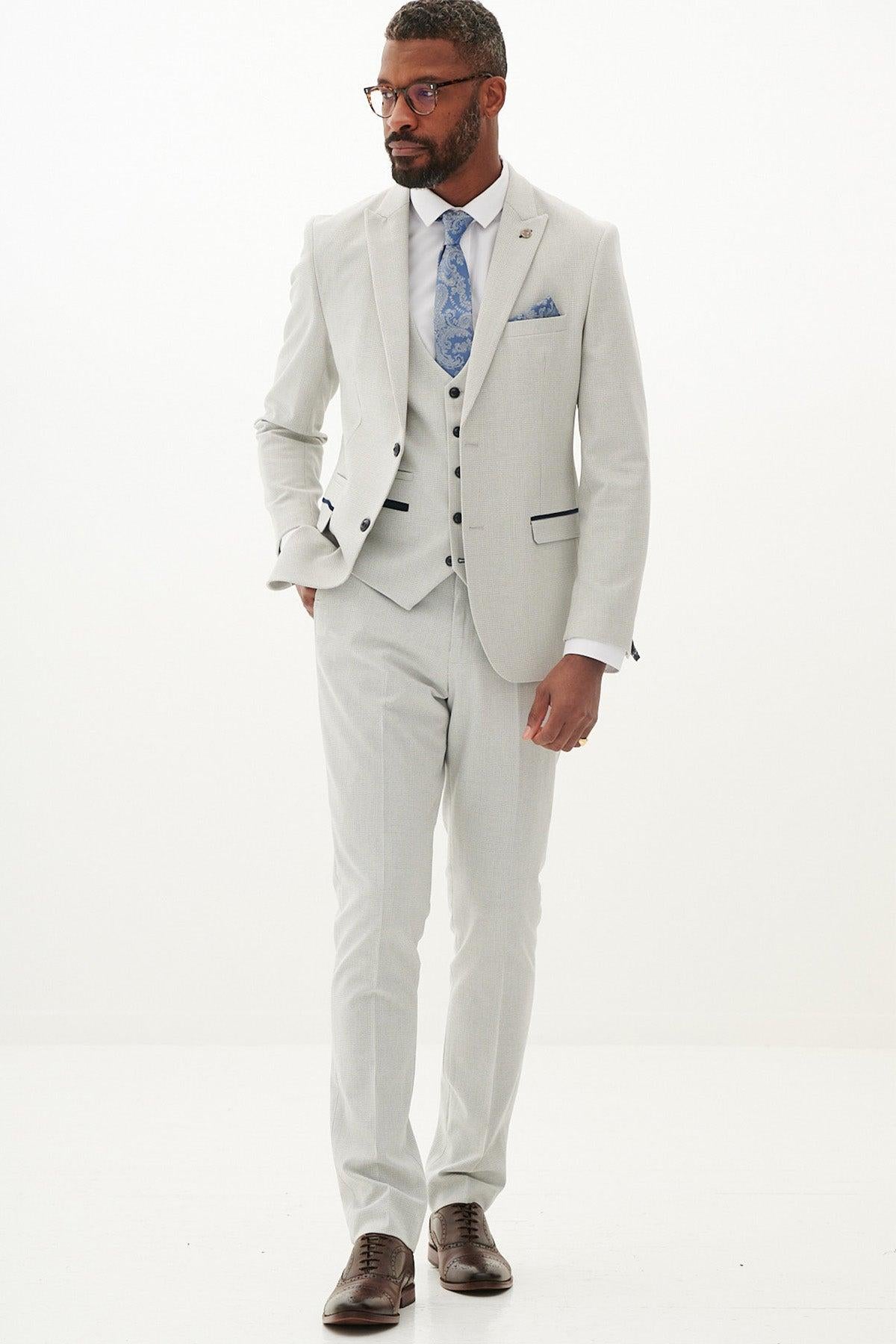 A man with short hair and glasses wears the Marc Darcy BROMLEY Stone Check Three Piece Suit, paired with a white shirt, blue patterned tie, pocket square, and brown dress shoes, standing against a plain white background.