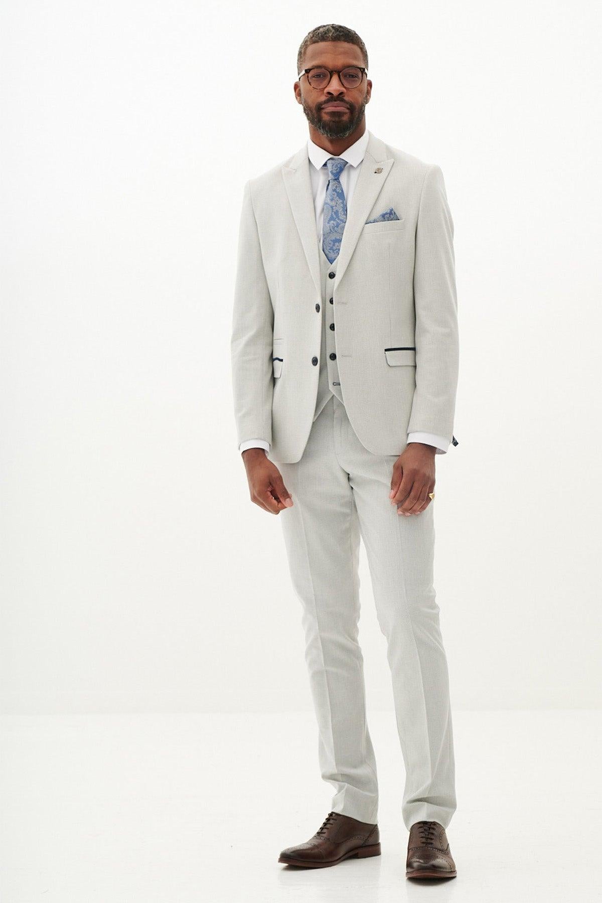 A man in the Marc Darcy BROMLEY - Stone Check Three Piece Suit with a blue patterned tie, white shirt, and vest stands against a plain white background. He wears glasses, has short hair, and brown dress shoes.