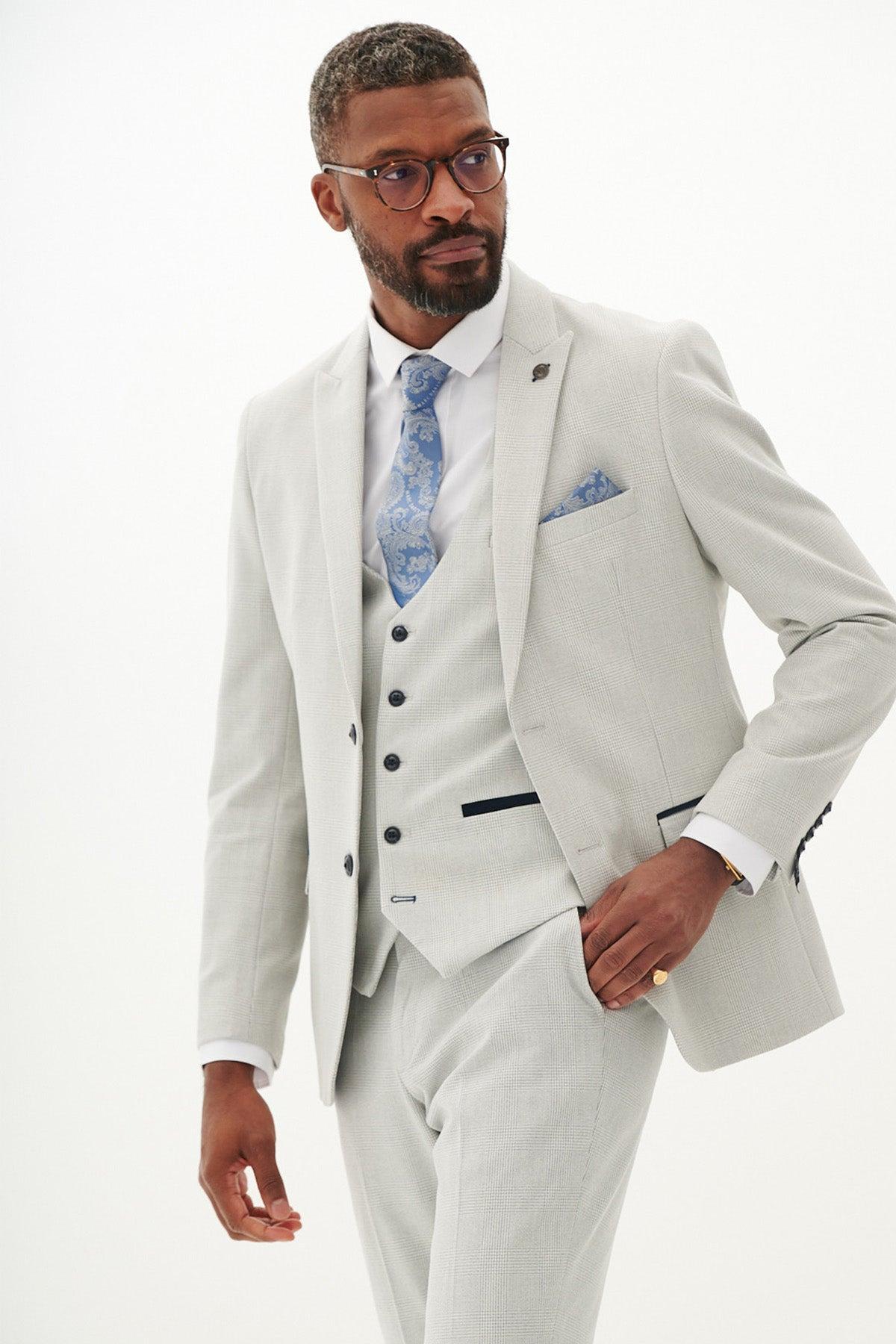 A man in the Marc Darcy BROMLEY stone check three piece suit, white shirt, blue paisley tie, and glasses stands with his hand in his pocket.