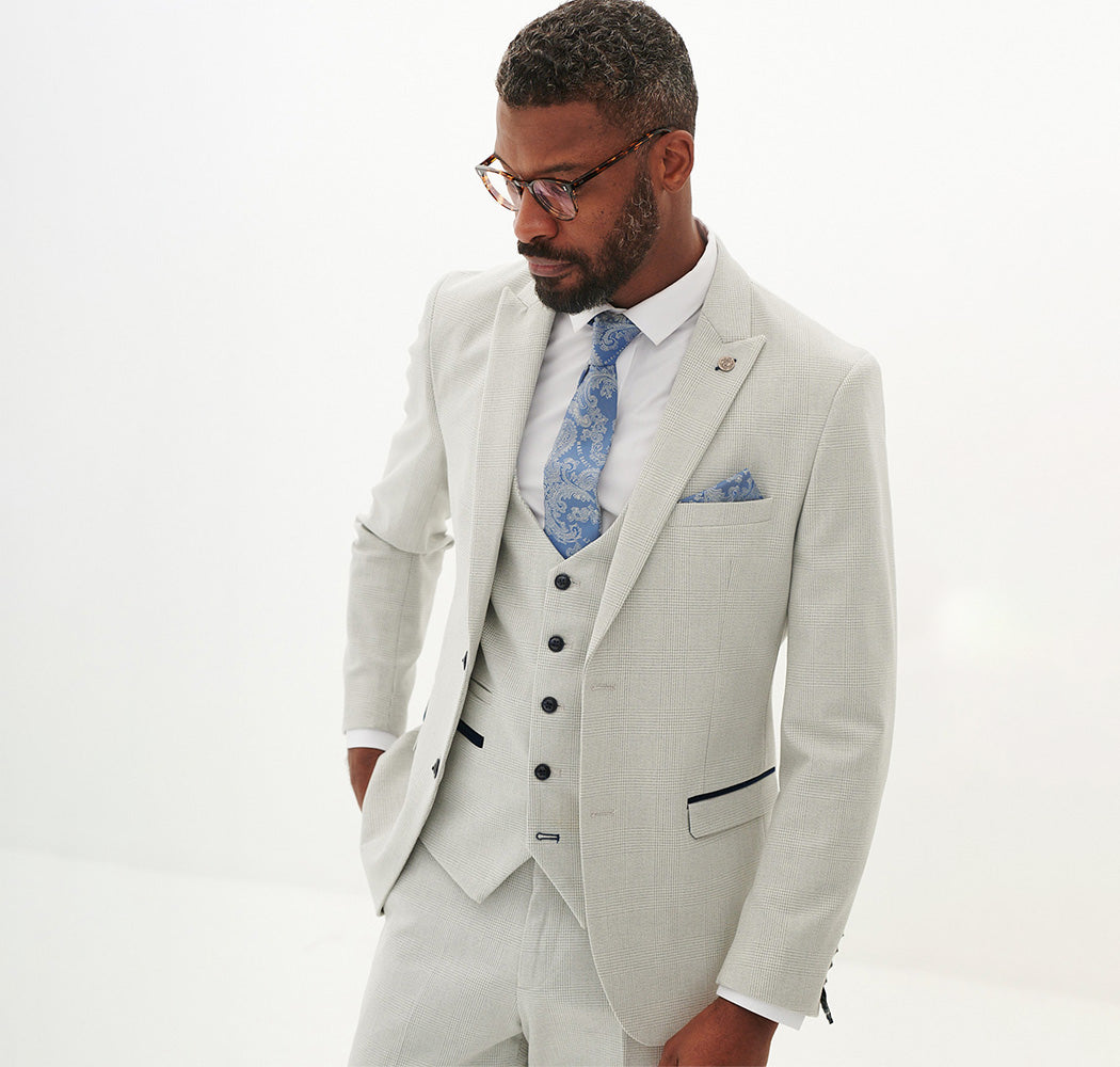 A man with short hair and a beard wears a light gray three-piece suit with a blue patterned tie and matching pocket square. He looks down and has one hand in his pocket against a plain white background.