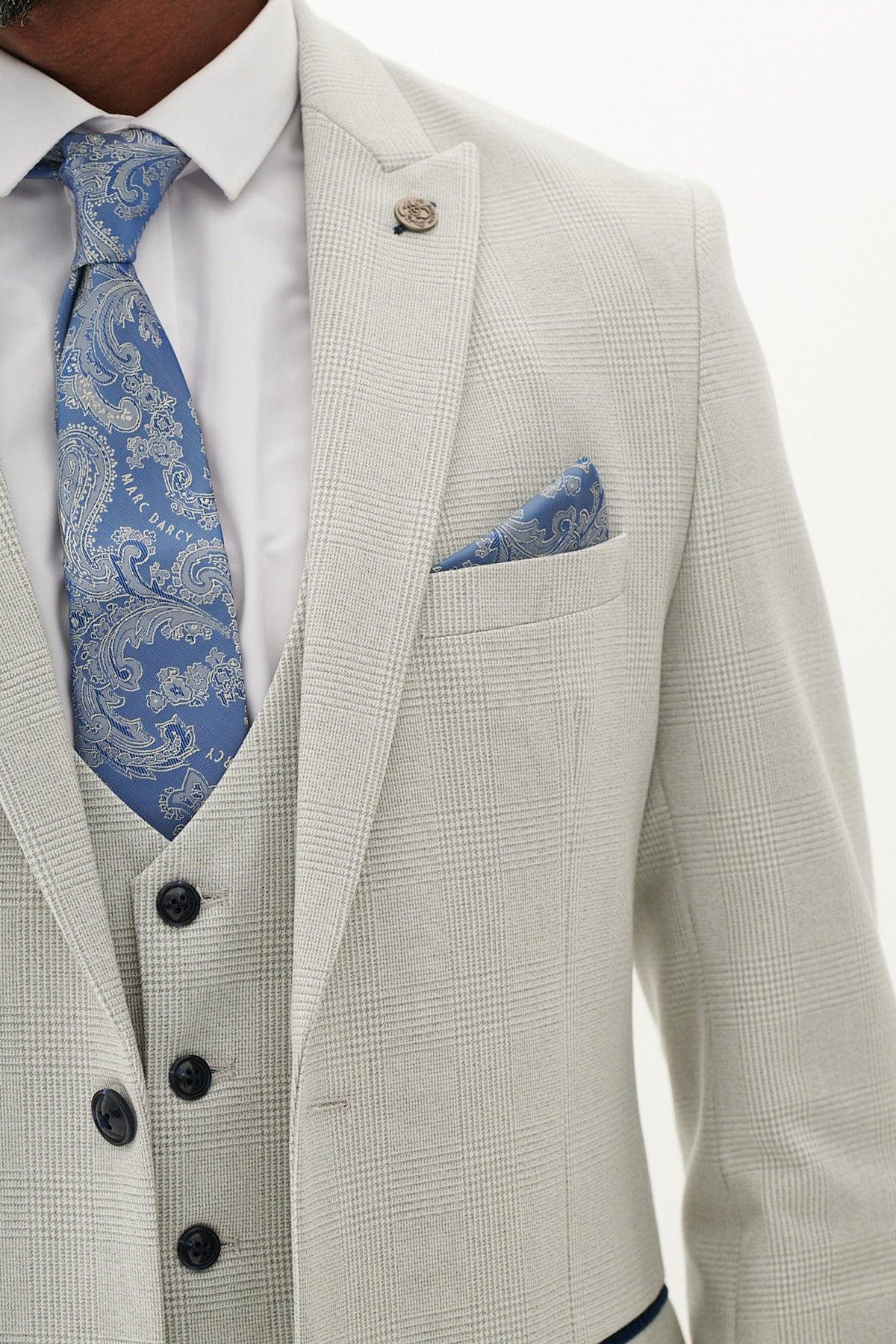 A close-up of a person wearing the Marc Darcy BROMLEY Stone Check Three Piece Suit, styled with a white shirt, blue paisley tie, matching pocket square, dark buttons, and lapel pin against a plain white background.