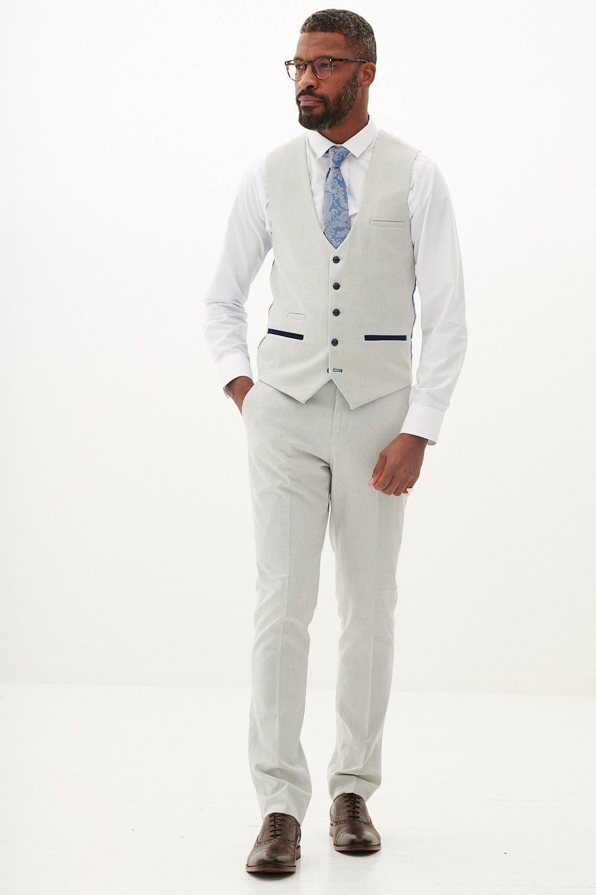 A man wears the Marc Darcy BROMLEY Stone Single Breasted Check Waistcoat with matching pants, white shirt, blue patterned tie, and brown shoes, standing against a white background with one hand in his pocket.