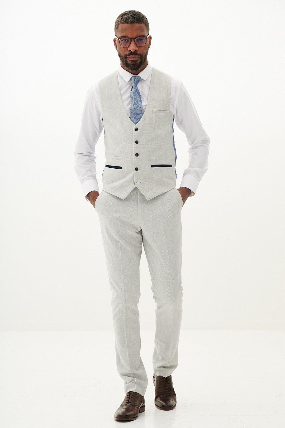 A man in a light gray suit with a white shirt and blue patterned tie wears glasses and completes his look with the Marc Darcy BROMLEY - Stone Single Breasted Check Waistcoat for added sophistication.
