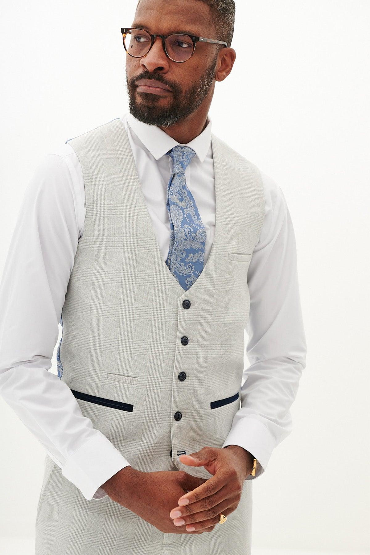A man in glasses, a white shirt, Marc Darcy’s BROMLEY Stone Single Breasted Check Waistcoat, and a blue patterned tie stands against a plain white background, looking off to the side.