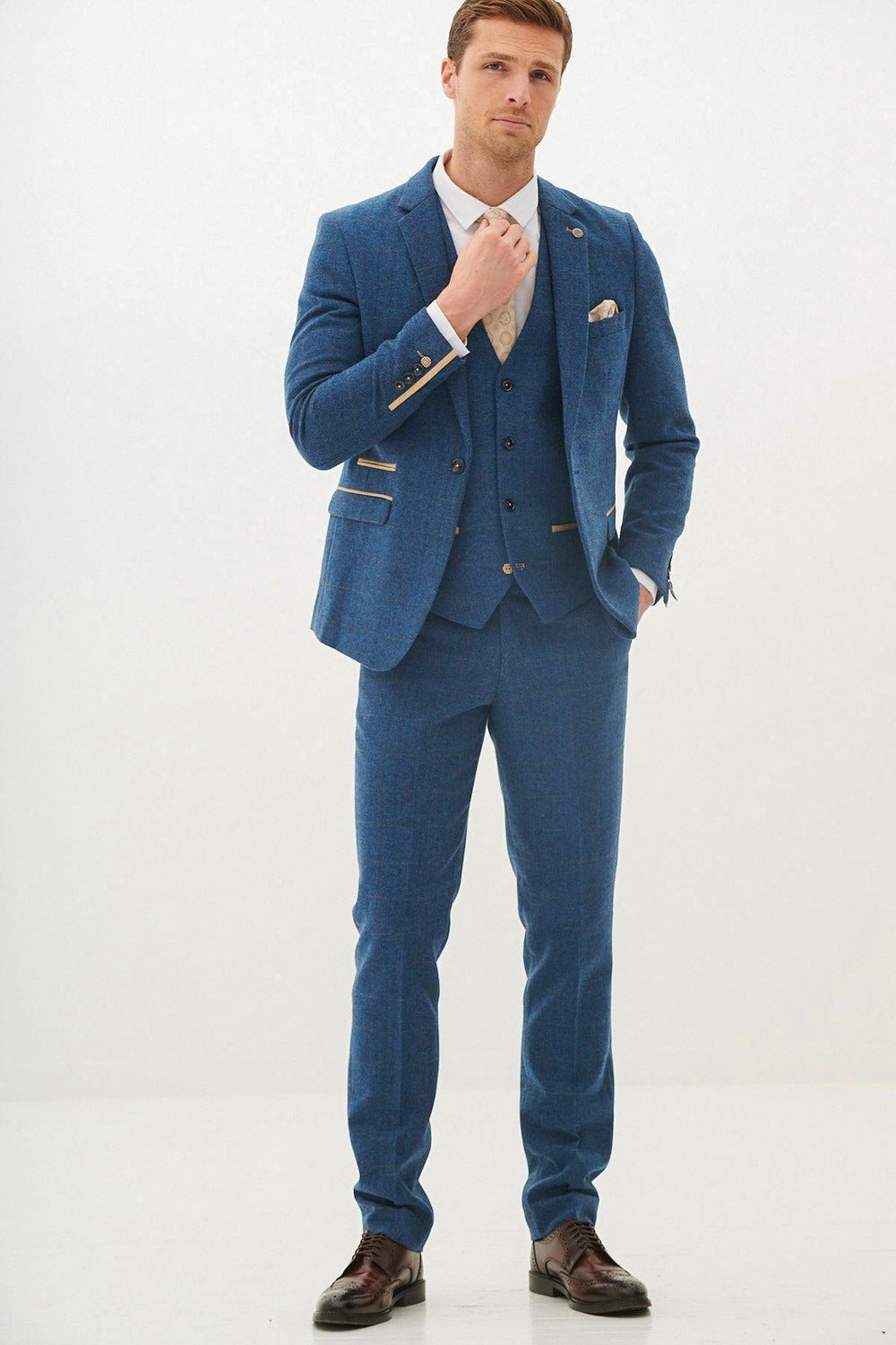 A man in the marcdarcy DION Blue Tweed Check Three Piece Suit with a white shirt and tan tie adjusts his tie against a white background.