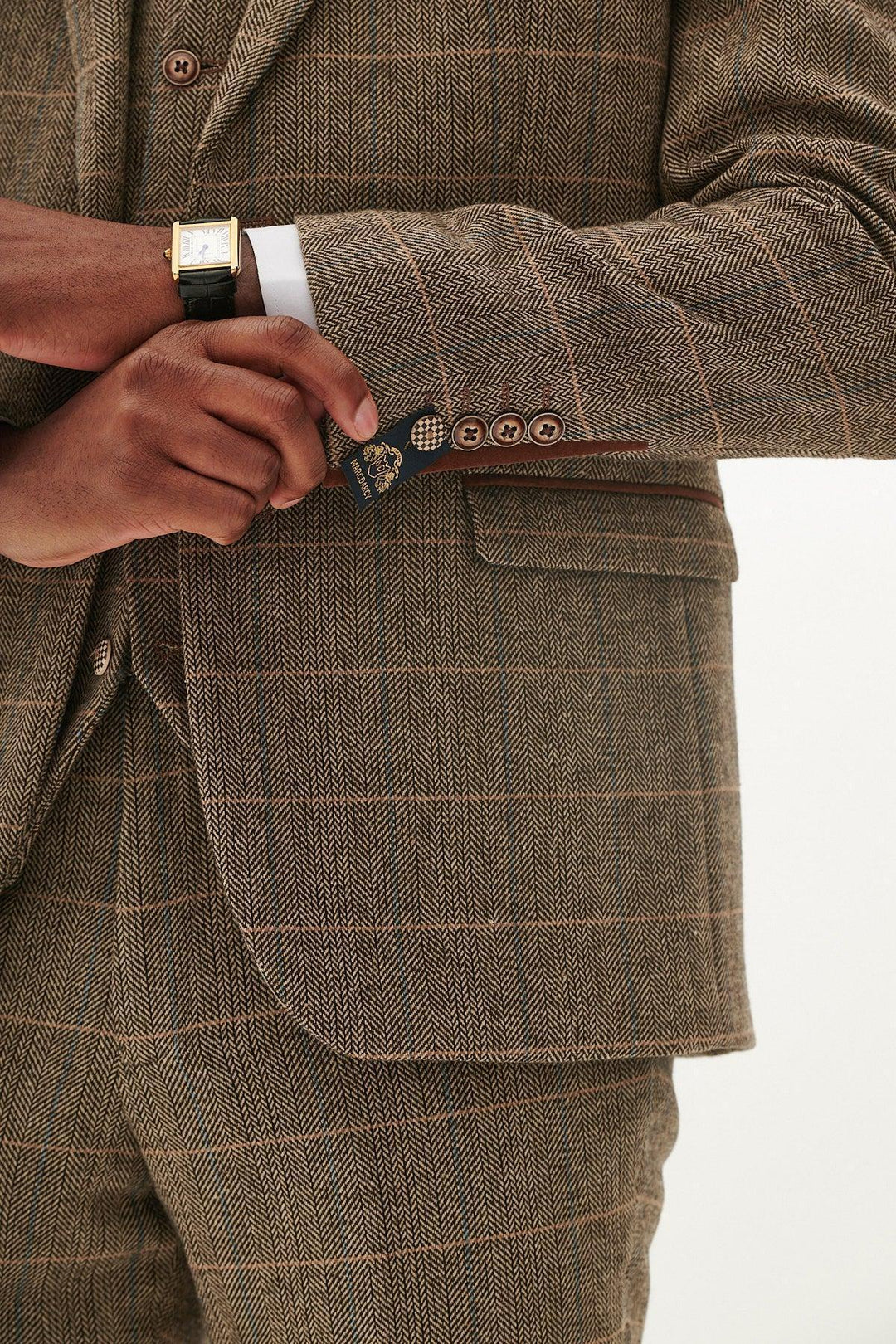 A person wearing the marcdarcy TED Tan Tweed Check Blazer adjusts their cuff, showing four brown buttons, a gold watch, and a blue tag. The blazer’s classic check pattern radiates timeless elegance.