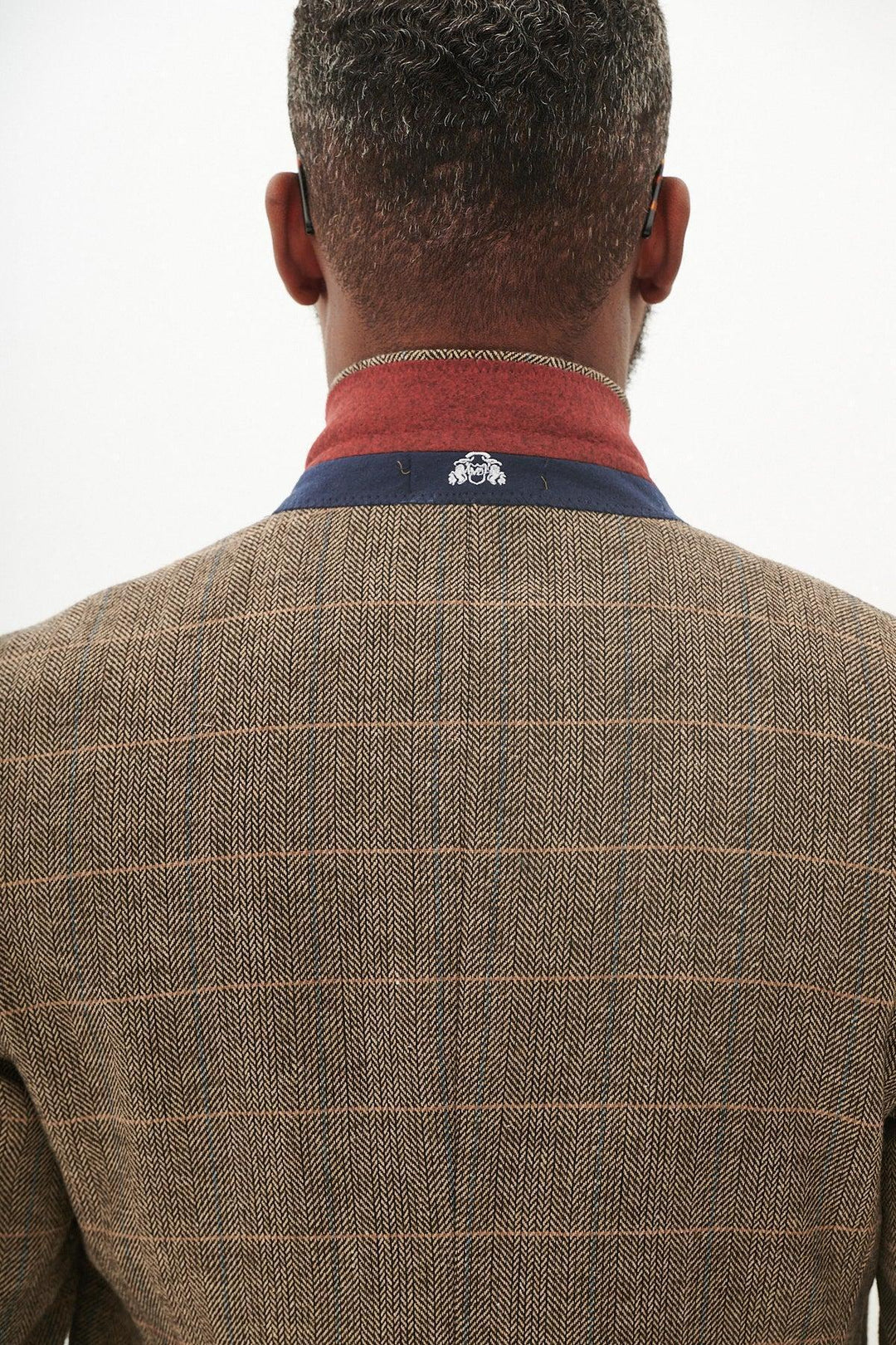 A man with short, curly hair is seen from behind wearing the marcdarcy TED - Tan Tweed Check Blazer, featuring a navy and red collar with a small white embroidered elephant logo on the back.