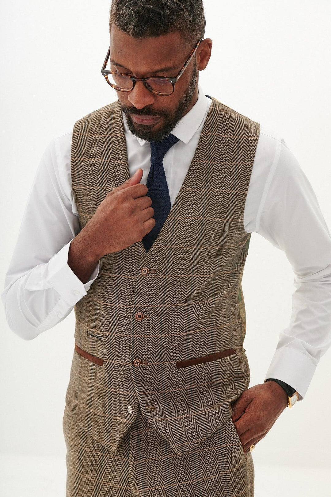 A man wearing glasses, a white shirt, blue tie, marcdarcy’s TED Tan Tweed Check Waistcoat with matching trousers stands against a plain light background, looking down and adjusting his vest—ideal for a country barn wedding.