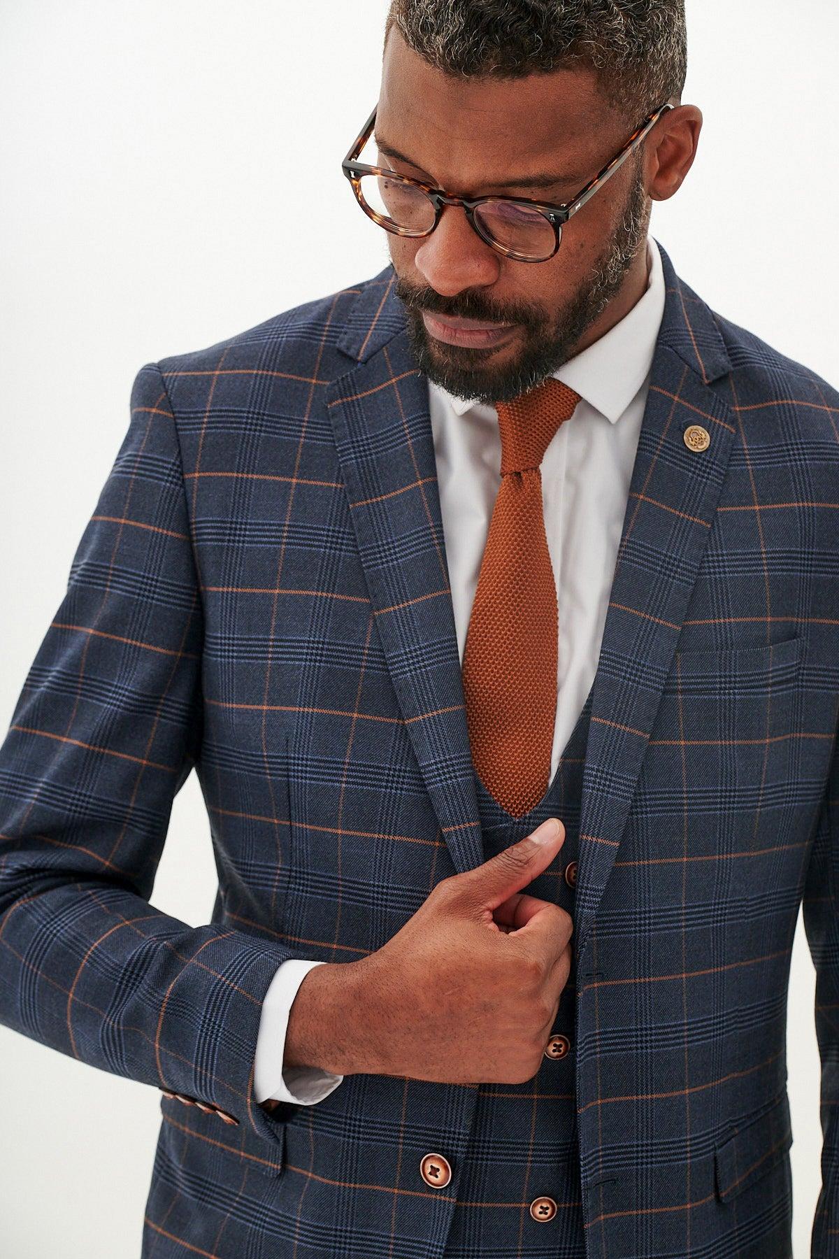 A man with short hair and a beard, wearing glasses, styles the marcdarcy JENSON - Marine Navy Check Blazer over a white dress shirt with a textured brown tie, adjusting his jacket—a smart suit choice for weddings.