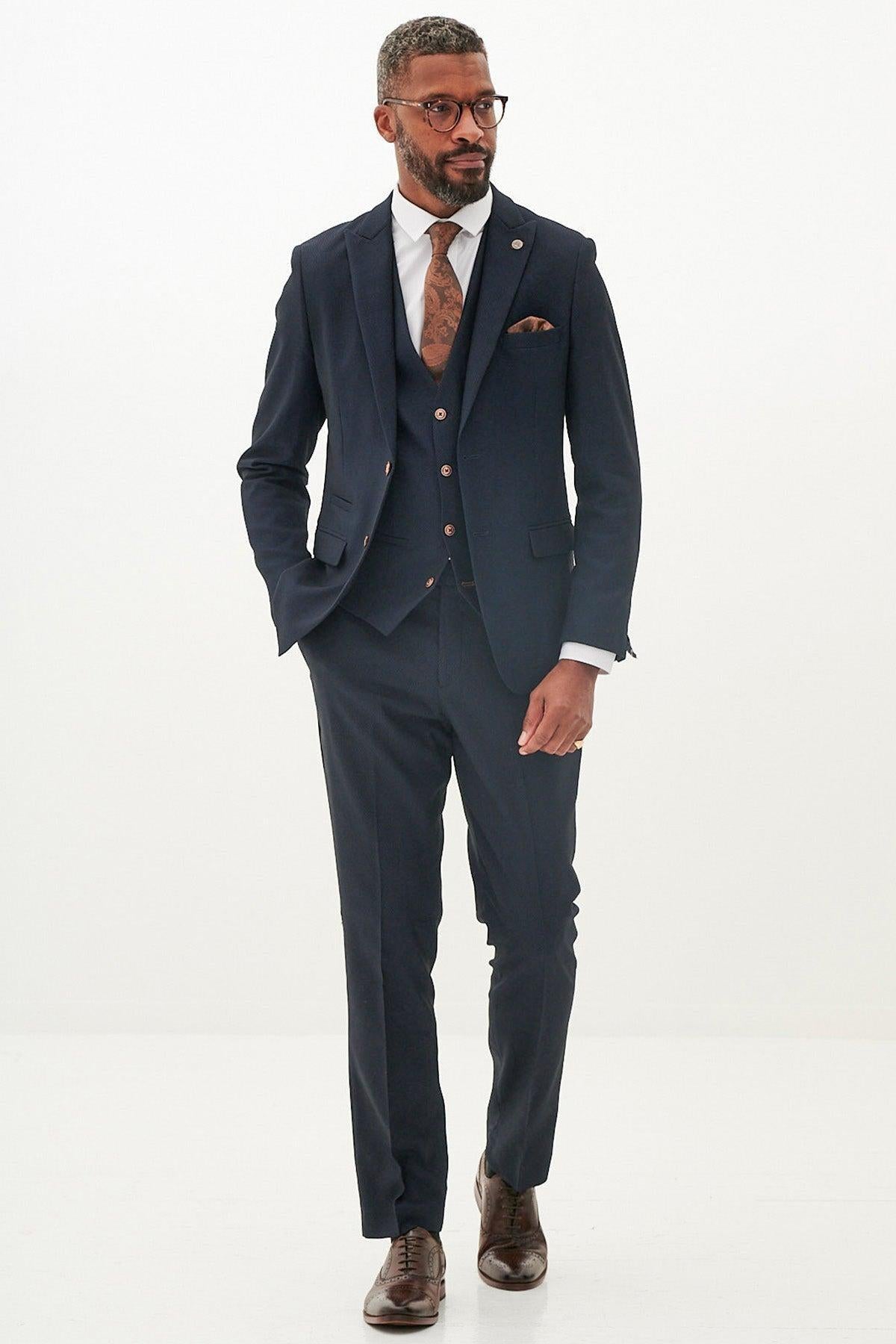 A man with glasses and a beard wears the marcdarcy MAX Navy Three Piece Suit with Contrast Buttons, paired with a white shirt, brown patterned tie, and brown shoes, standing hands-in-pockets against a plain white background.