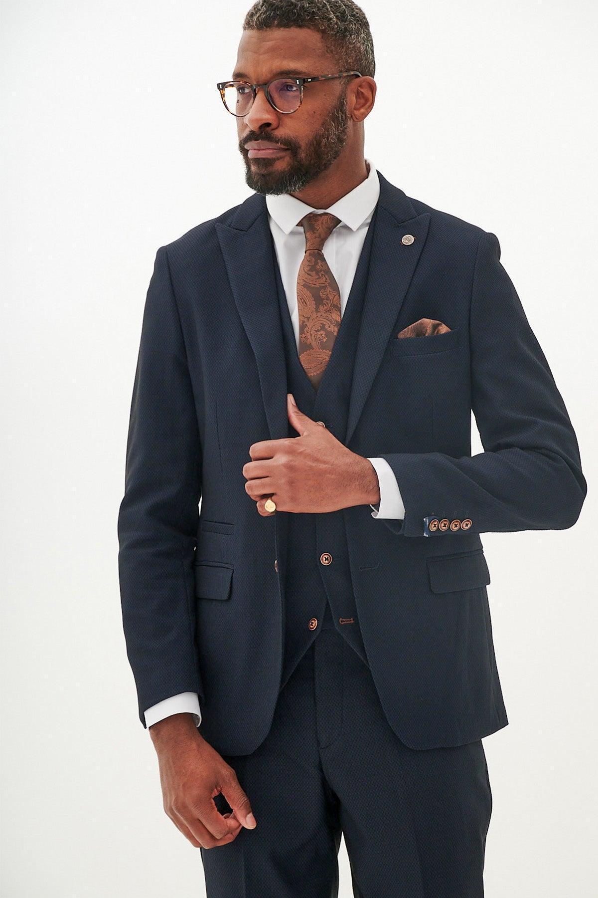 A man in the marcdarcy MAX Navy Three Piece Suit with contrast buttons stands against a plain white background, wearing glasses, a brown patterned tie, and matching pocket square, looking slightly to the side.