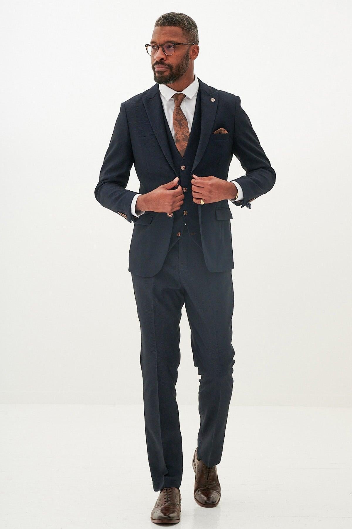 A man in the marcdarcy MAX Navy Three Piece Suit with Contrast Buttons, white shirt, patterned tie, and brown shoes adjusts his peak lapel jacket while looking to the side against a plain white background. He has short hair and glasses.