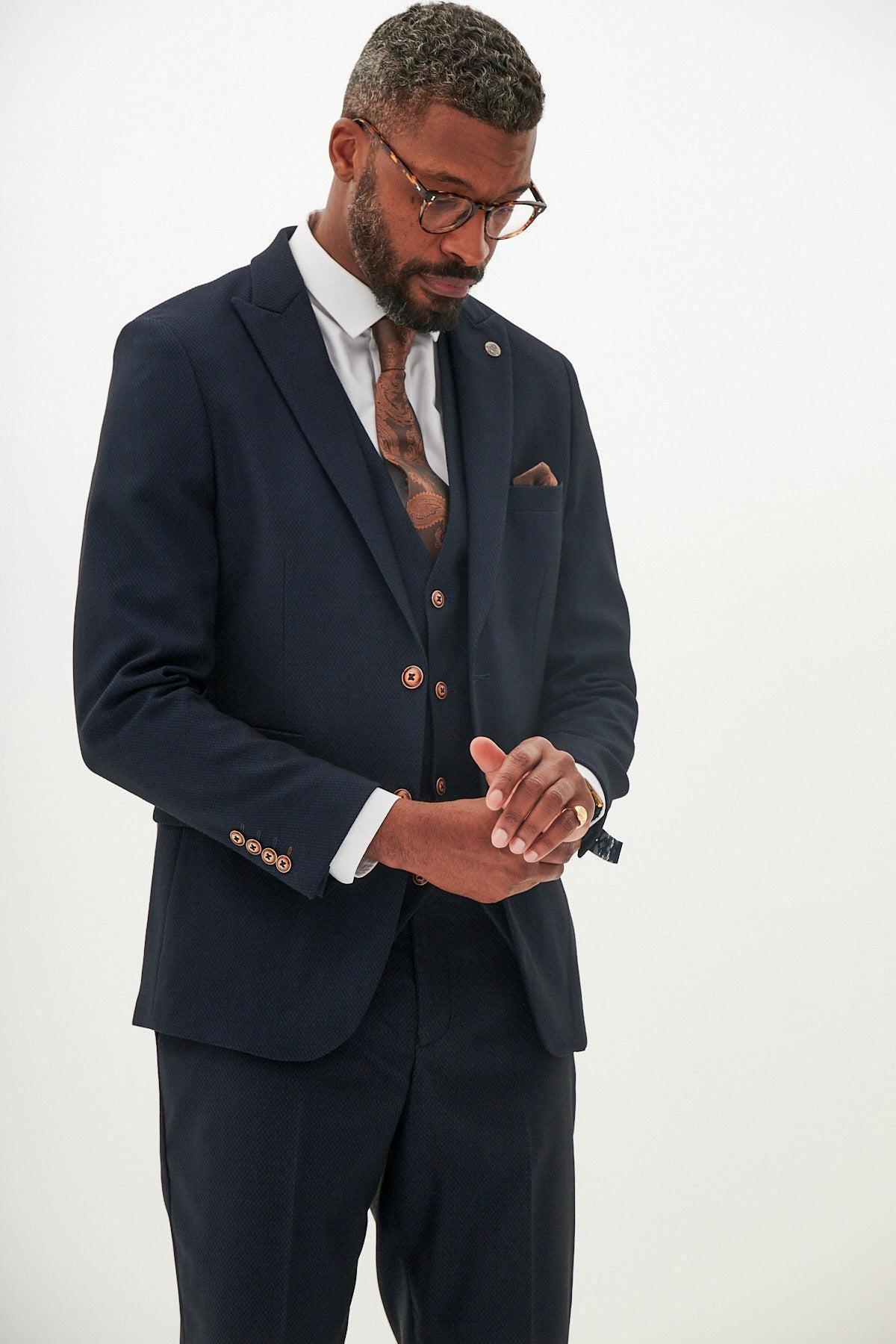 A man wearing the marcdarcy MAX Navy Three Piece Suit with contrast buttons, paired with a white shirt and patterned tie, adjusts his watch. He has glasses, short hair, a beard, and visible tattoos on his chest and hands.
