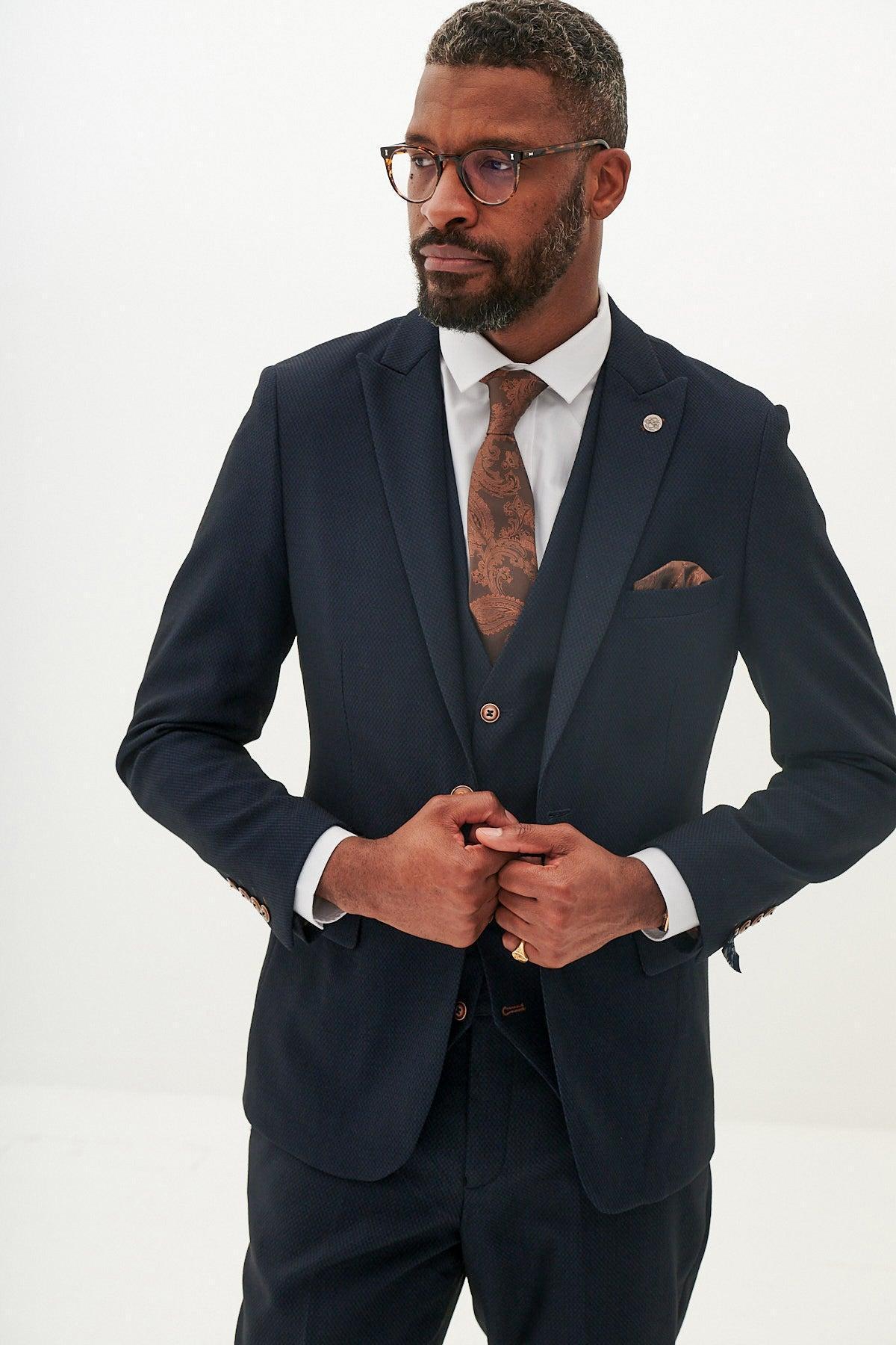 A man with short hair, glasses, and a beard wears the marcdarcy MAX Navy Three Piece Suit with contrast buttons, paired with a white shirt and patterned brown tie, adjusting his jacket against a plain white background.