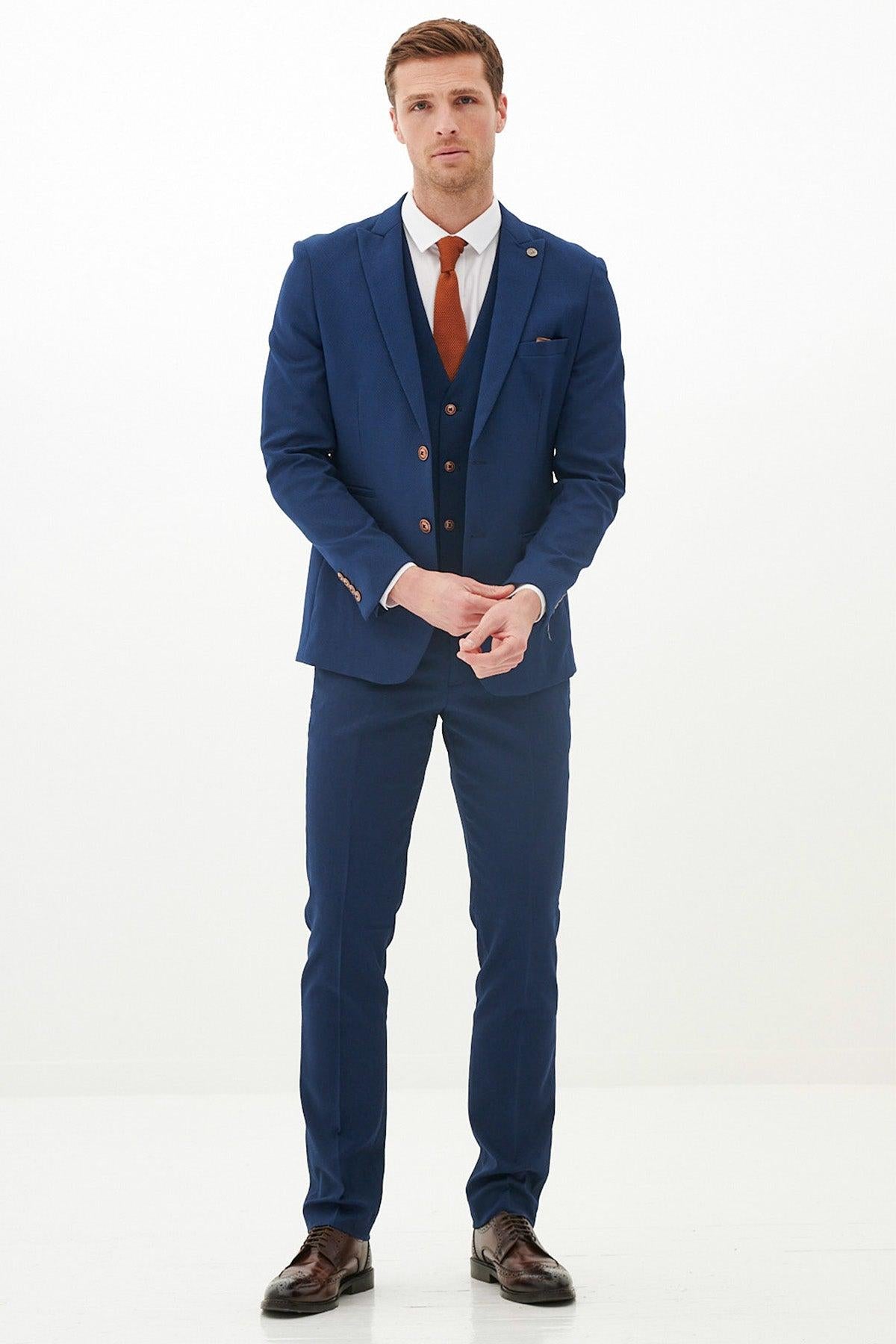 A man stands against a plain white background wearing the marcdarcy MAX - Royal Blue Three Piece Suit With Contrast Buttons, paired with a white shirt, orange tie, and pocket square. He has short hair and looks forward with his hands clasped.