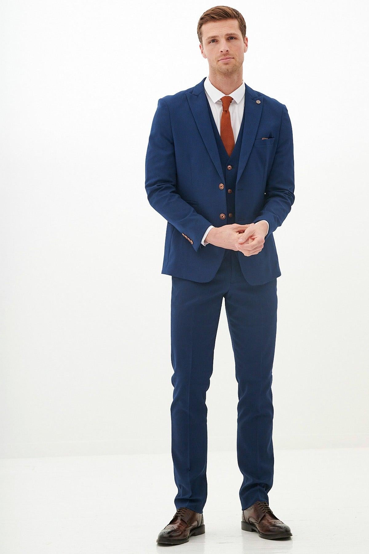 A man stands against a white background wearing the marcdarcy MAX - Royal Blue Three Piece Suit With Contrast Buttons, paired with a white dress shirt, brown tie, and brown dress shoes. He clasps his hands and looks at the camera.