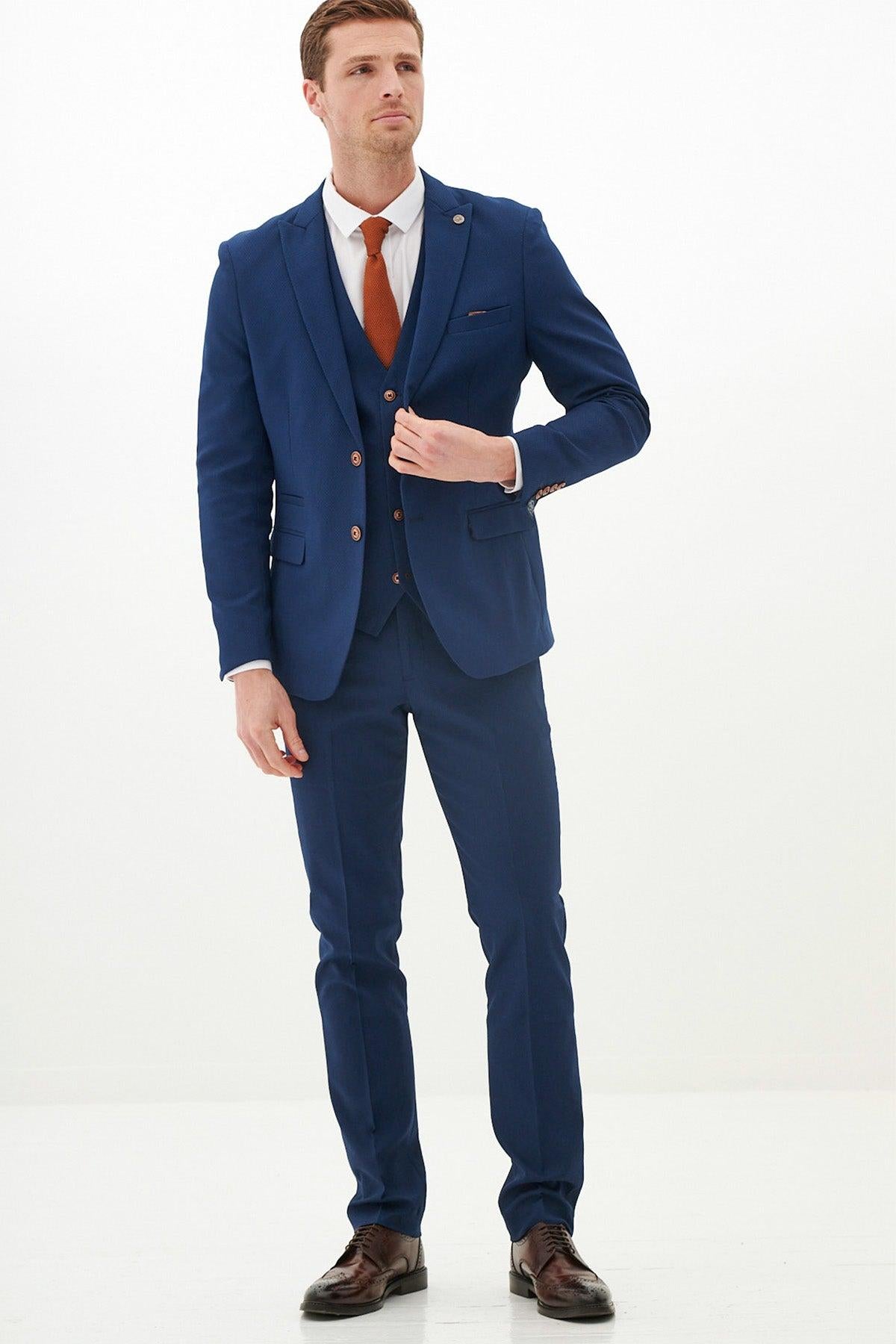 A man wears the marcdarcy MAX Royal Blue Three Piece Suit With Contrast Buttons, paired with a white shirt and rust-colored tie, standing against a white background.