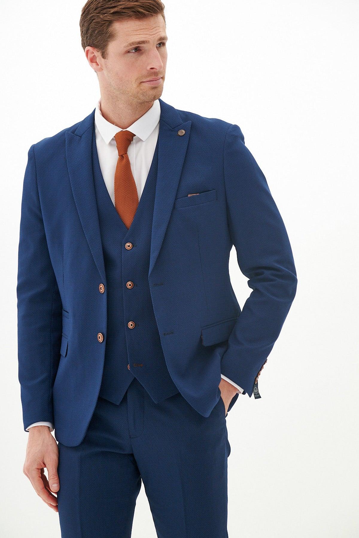 A man wears the marcdarcy MAX - Royal Blue Three Piece Suit With Contrast Buttons, including a slim fit blazer, white shirt, and orange tie, standing with his hand in his pocket against a white background.
