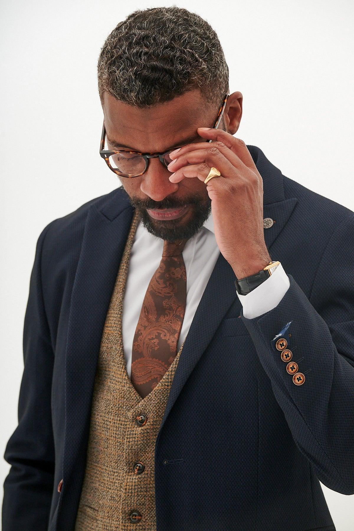 A man in the marcdarcy MAX - Navy Suit with Kris Tan Tweed Waistcoat, styled with a brown tie, watch, and ring, adjusts his glasses while looking down—a sharp formal look set against a plain background.