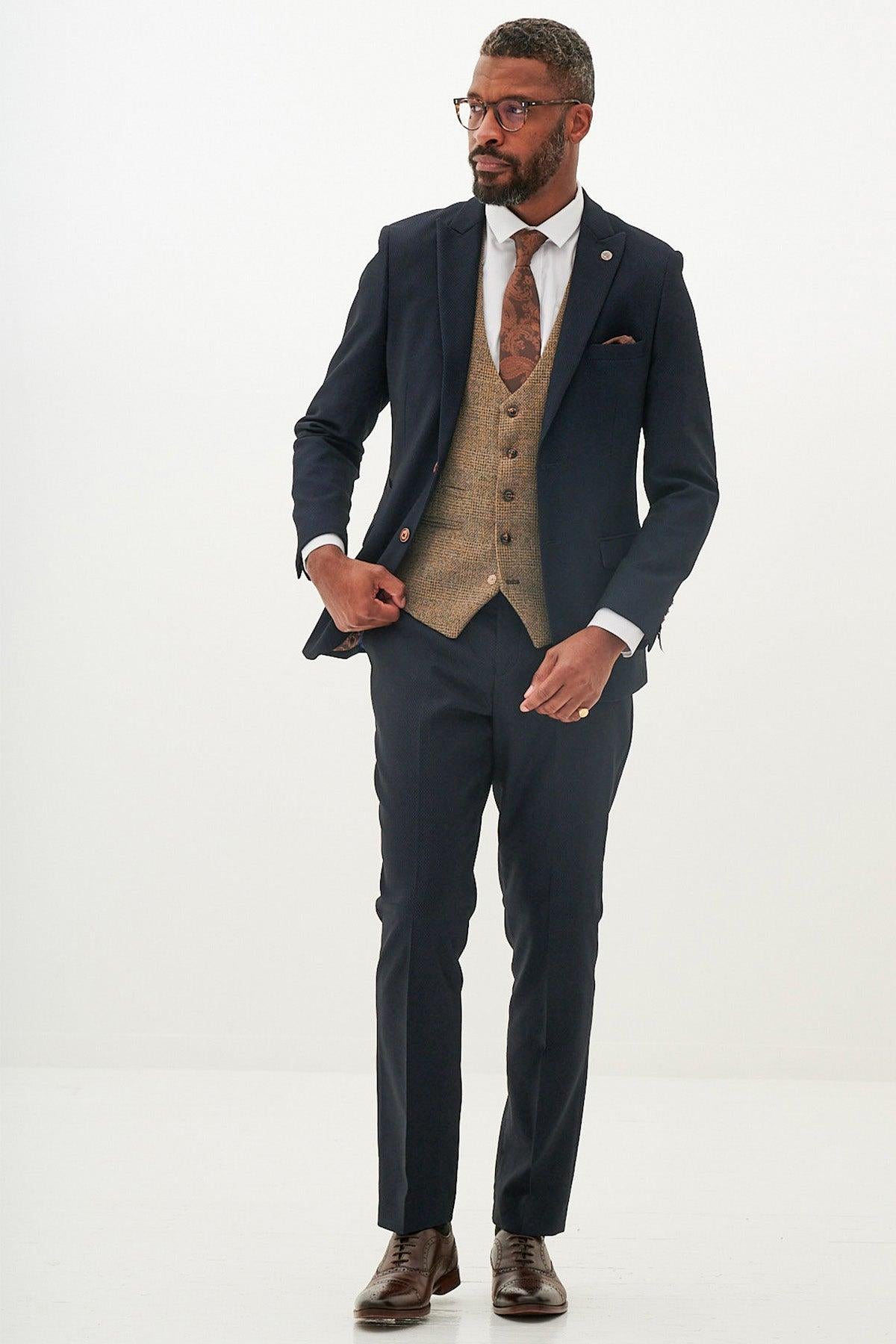 A man wears the marcdarcy MAX - Navy Suit with Kris Tan Tweed Waistcoat and a brown patterned tie, posing against a white background—a refined formal look.