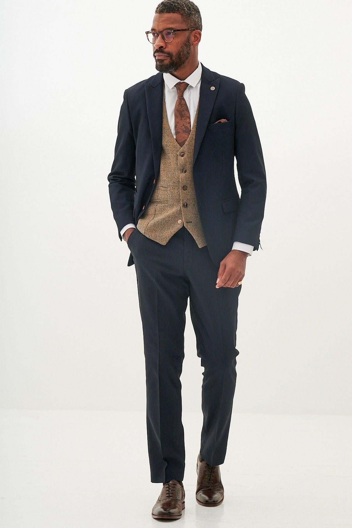A man wears the marcdarcy MAX Navy Suit with Kris Tan Tweed Waistcoat, paired with a white shirt, patterned tie, glasses, and brown dress shoes for a stylish mixed look.