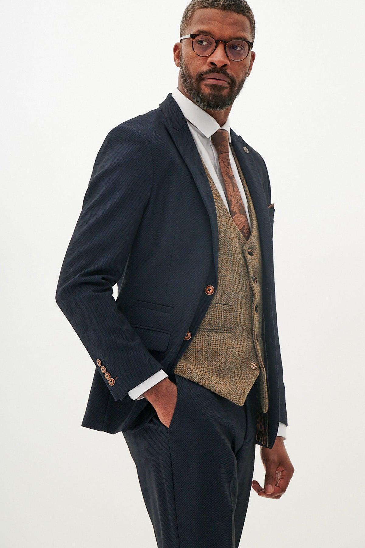 A man in a marcdarcy MAX Navy Suit with Kris Tan Tweed Waistcoat, styled with a white shirt and brown patterned tie, stands confidently with one hand in his pocket, gazing sideways against a plain backdrop.