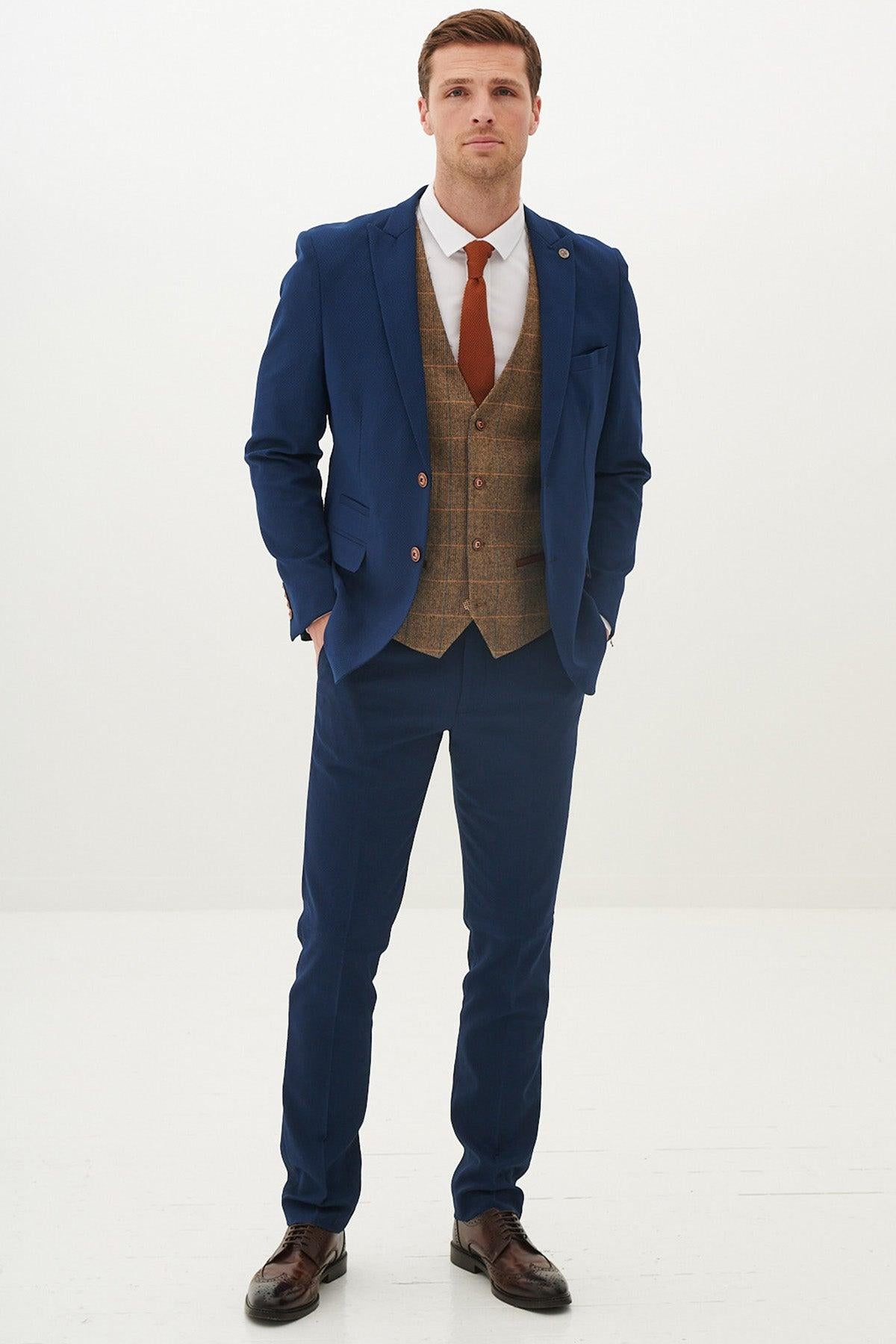 A man faces forward against a white background, wearing the marcdarcy MAX - Royal Blue Suit with Kris Tan Tweed Waistcoat, paired with a white shirt, rust tie, and brown dress shoes.