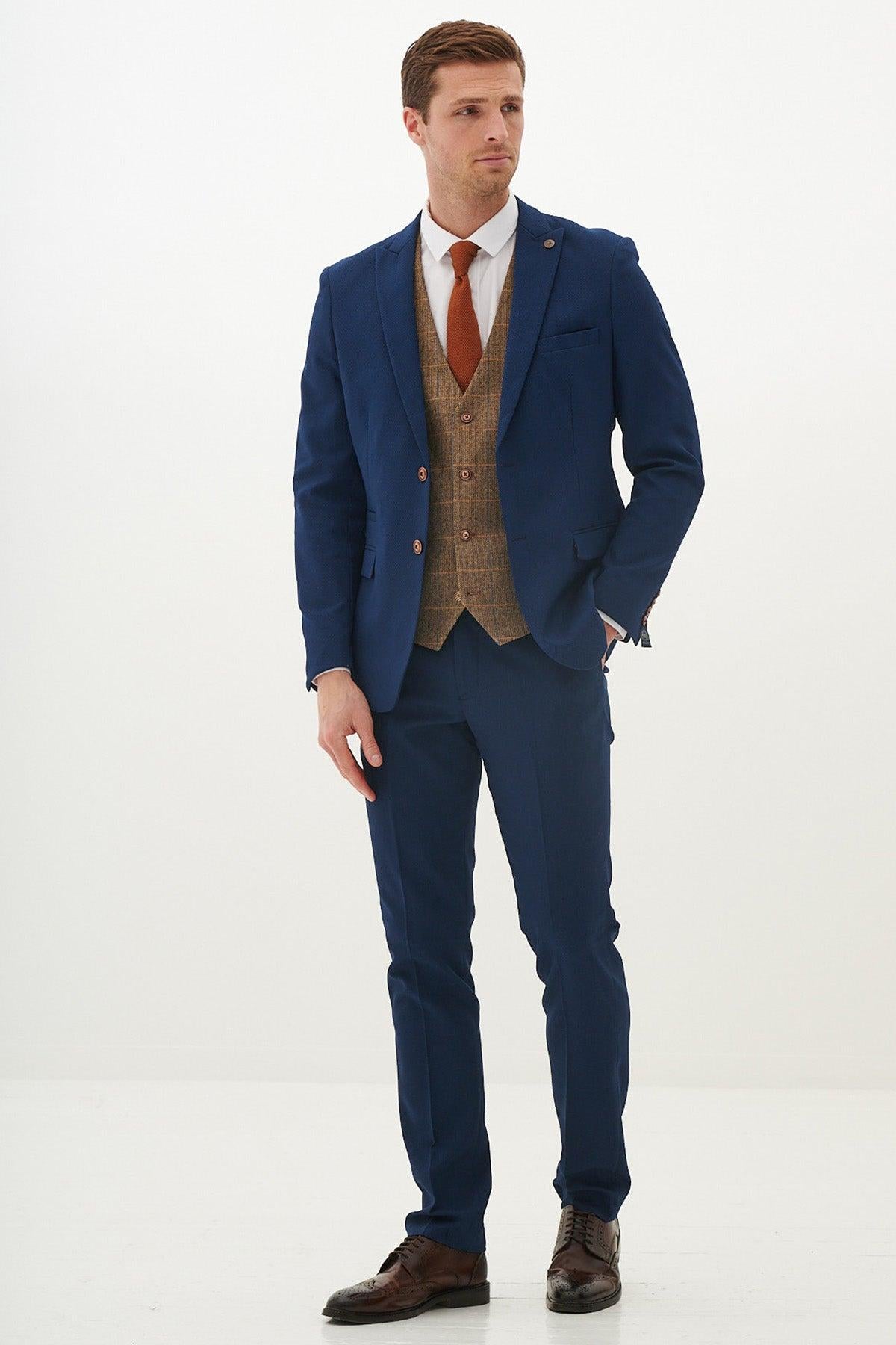 A man wearing the marcdarcy MAX - Royal Blue Suit With Kris Tan Tweed Suit stands against a white background, styled with a white shirt, orange tie, and brown dress shoes. He has short brown hair and a neutral expression, one hand in his pocket.
