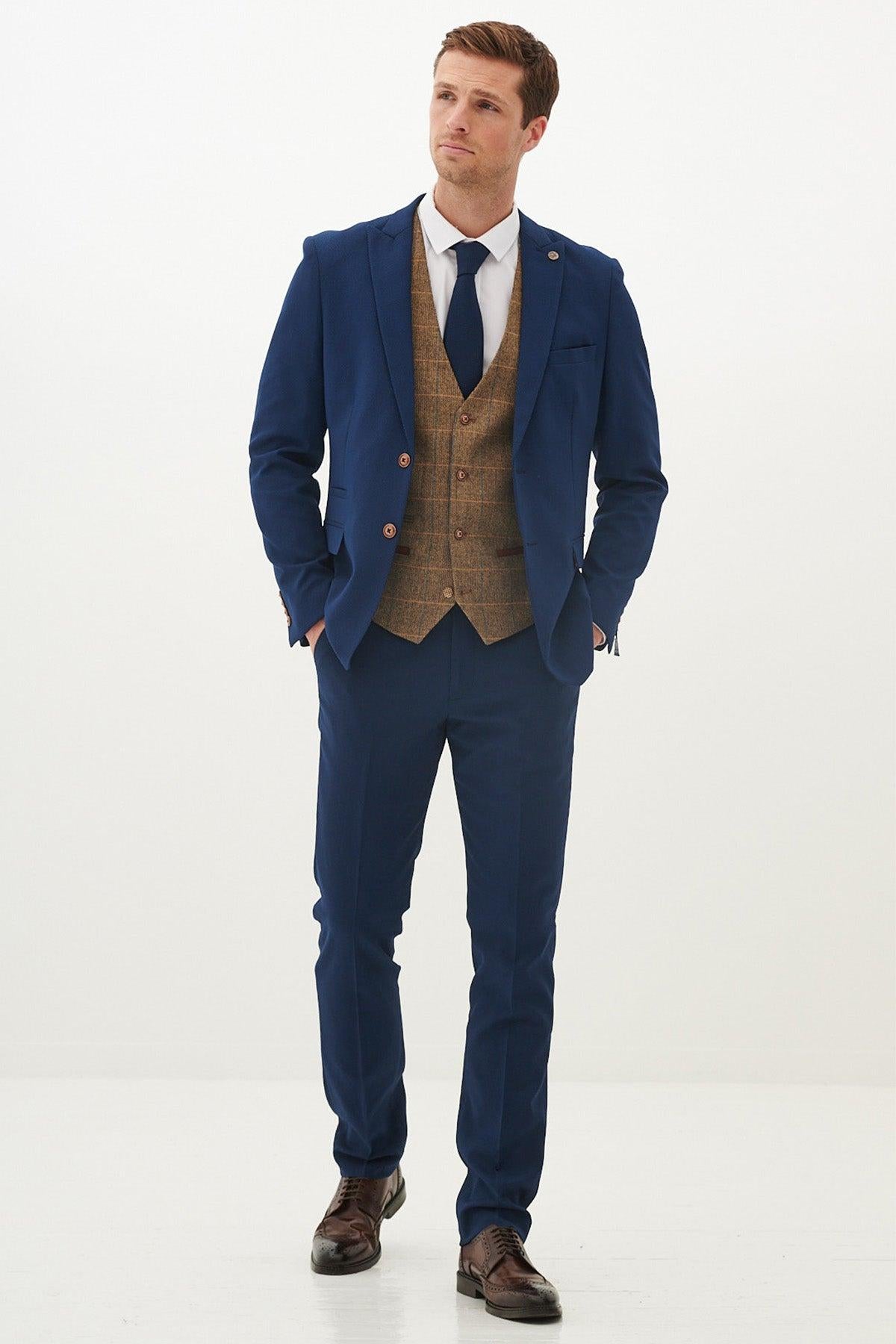 A man wears the marcdarcy MAX Royal Blue Suit with Kris Tan Tweed waistcoat, paired with a white shirt and navy tie, hands in his pockets.