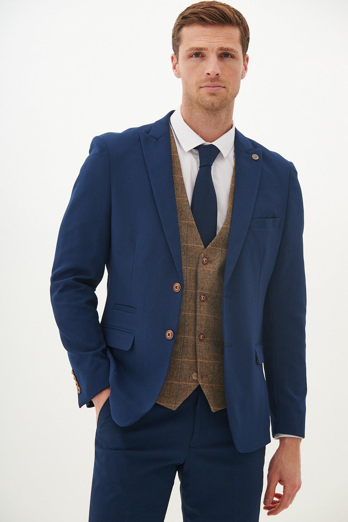 A man wearing the marcdarcy MAX Royal Blue Suit with Kris Tan Tweed Waistcoat stands against a plain background, one hand in his pocket, looking directly at the camera with a neutral expression.