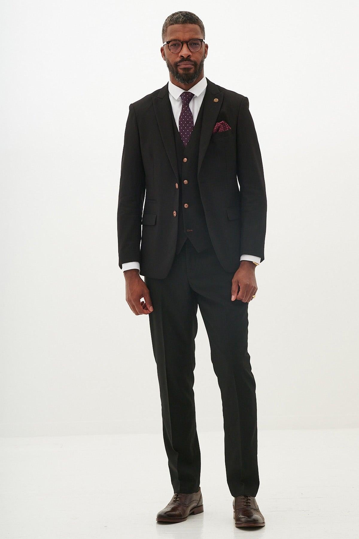 A man with short hair, beard, and glasses wears the marcdarcy MAX Black Three Piece Suit with Contrast Buttons, paired with a white shirt, patterned tie, and pocket square, standing against a plain white background.