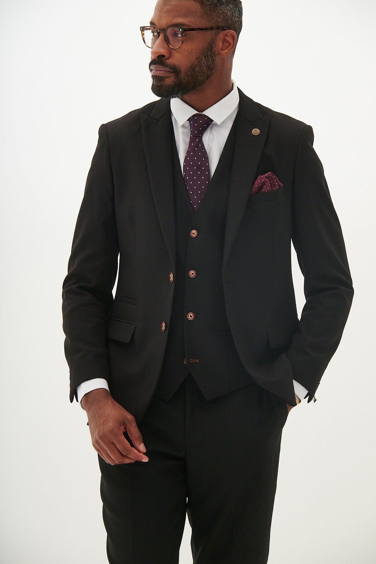 A man in the marcdarcy MAX black three piece suit with contrast buttons, styled with a polka dot tie, camouflage pocket square, and glasses, stands against a white background looking to the side with one hand at his waist.