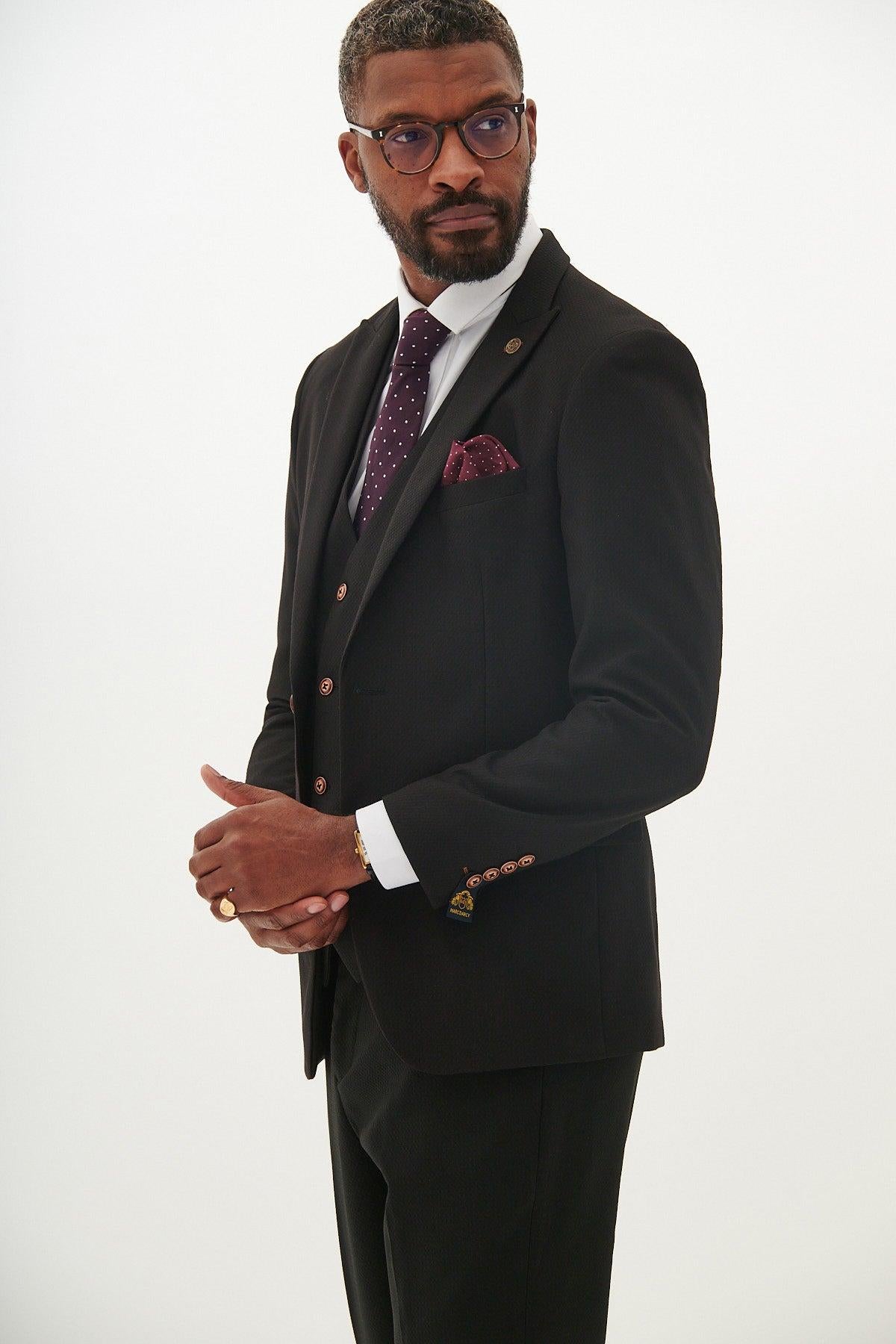 A man in a marcdarcy MAX black three piece slim-fit suit with contrast buttons, styled with a burgundy polka dot tie and matching pocket square, stands against a white background. He wears glasses, has short hair and a beard, looking to the side.