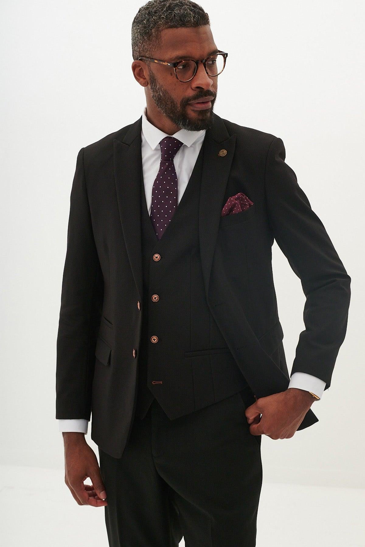A man in the marcdarcy MAX Black Three Piece Suit with contrast buttons, paired with a white shirt and purple polka dot tie, accessorized with a burgundy pocket square. He also wears glasses and has a beard.