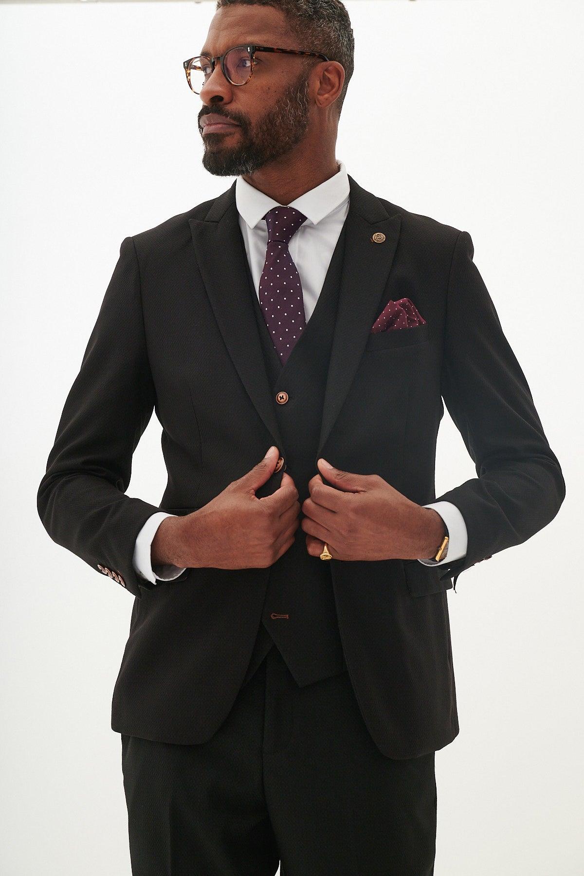 A man wears the marcdarcy MAX black three piece suit with contrast buttons, paired with a white shirt, purple polka-dot tie, pocket square, and glasses—modern workwear at its best against a plain white background.