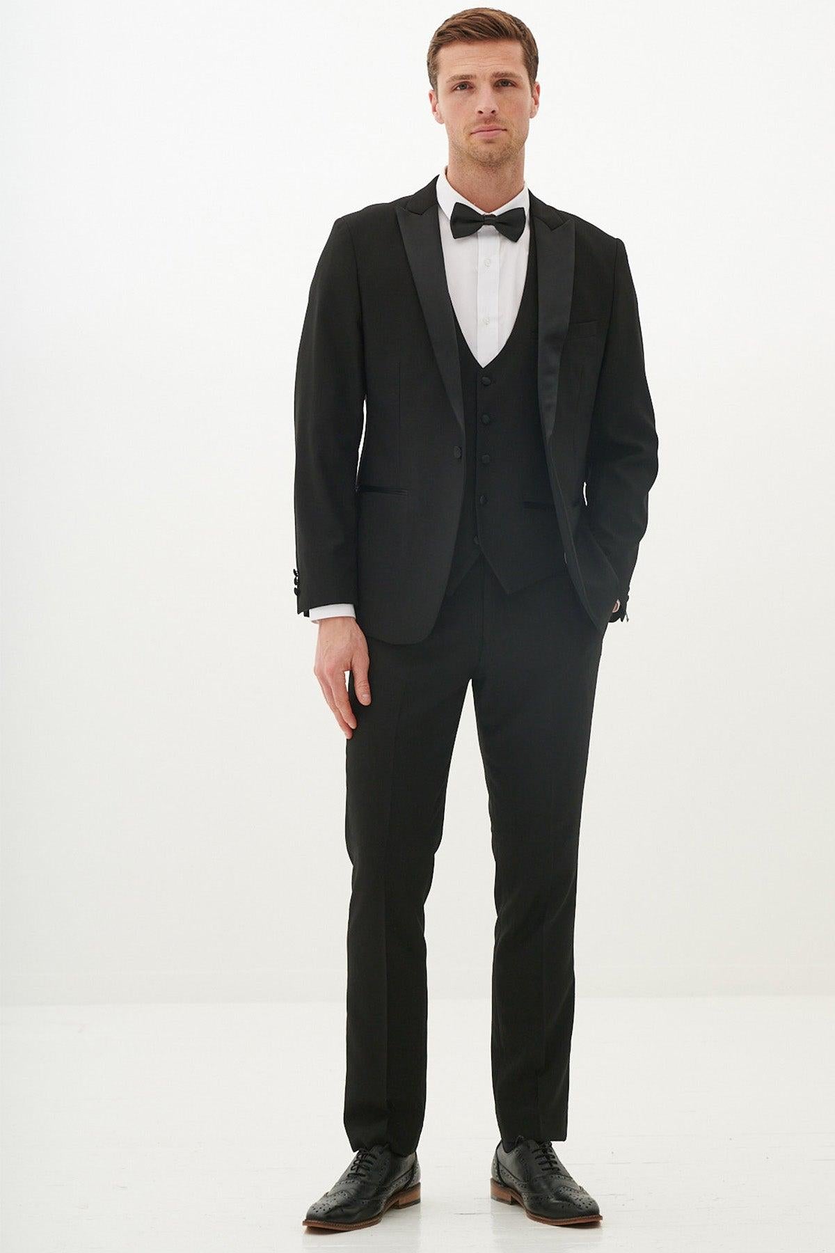 A man in the marcdarcy DALTON Black Tux Lapel Diamond Three Piece Suit with a white shirt and black bow tie stands against a white background.