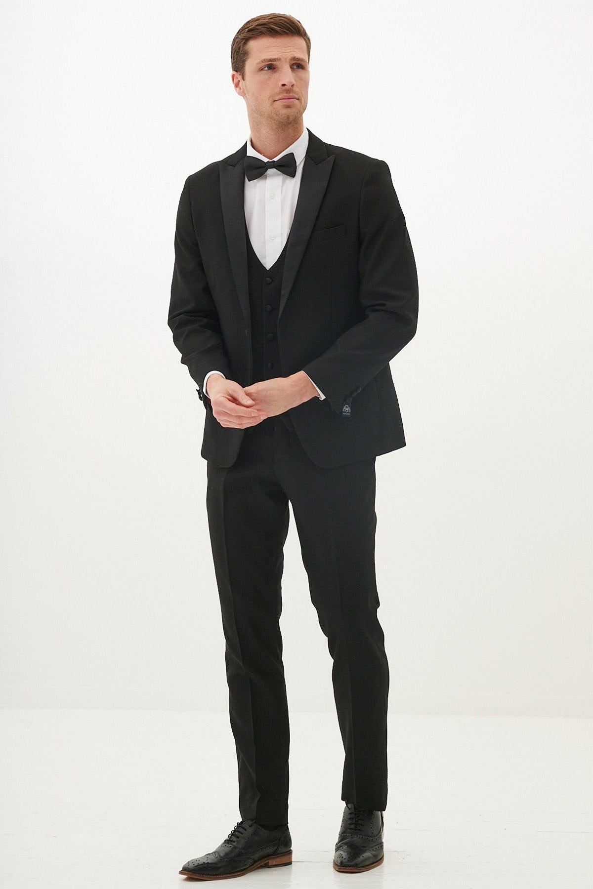 A man wears the marcdarcy DALTON Black Tux Lapel Diamond Three Piece Suit with a slim fit blazer, white shirt, and black bow tie, standing against a white background.