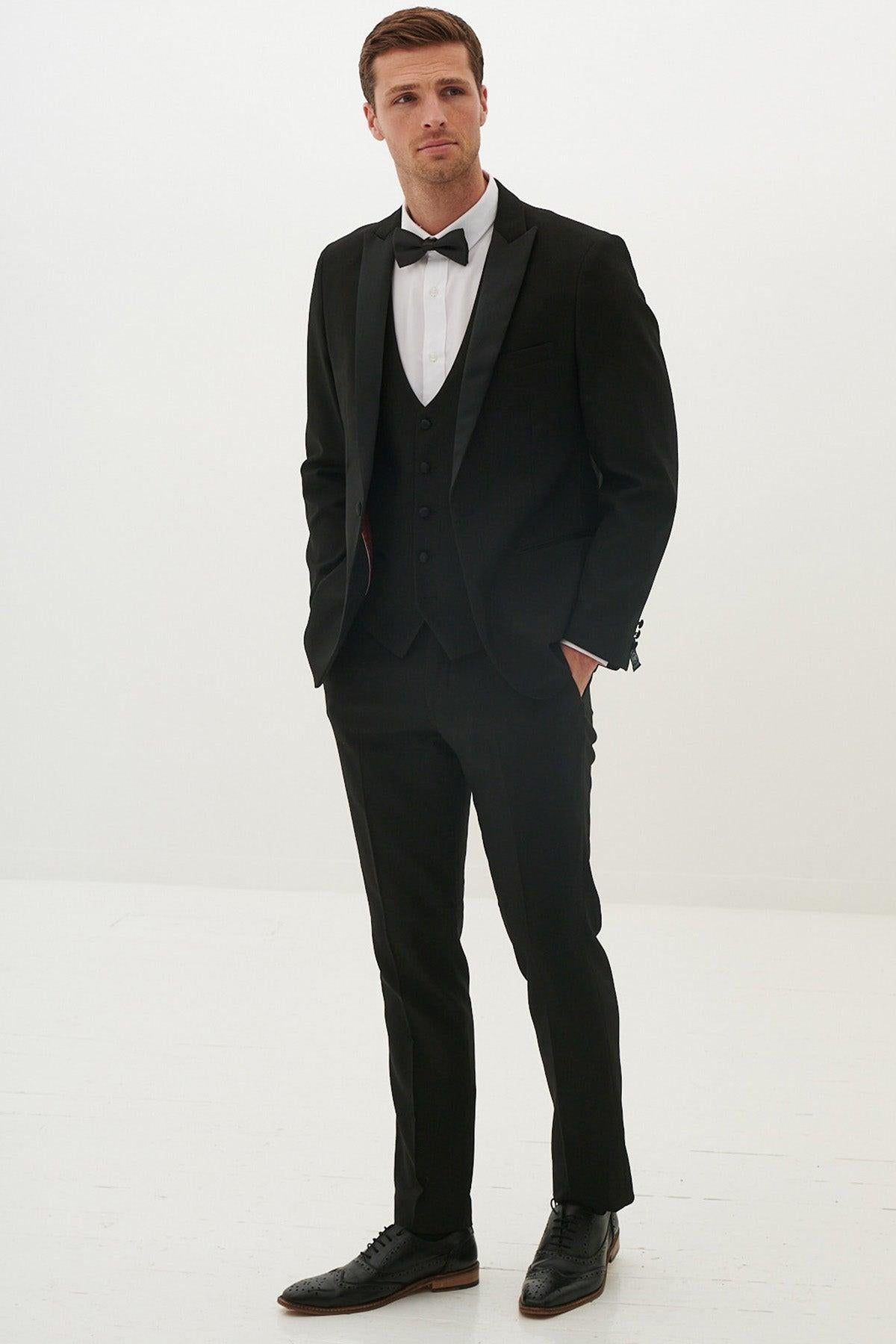 A man stands against a plain white background wearing the marcdarcy DALTON Black Tux Lapel Diamond Three Piece Suit, paired with a bow tie, white dress shirt, and black dress shoes, his hands casually in his pockets.